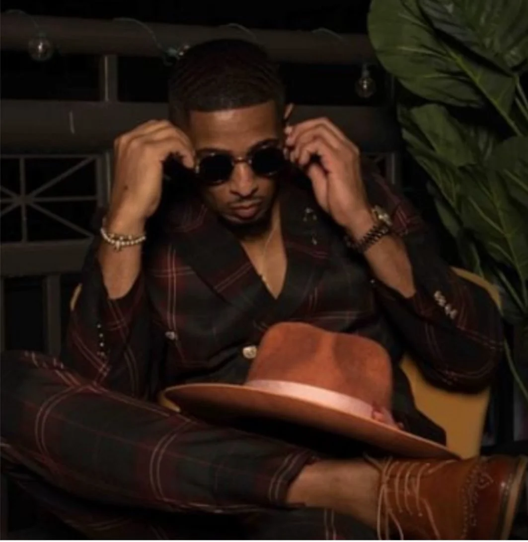 A man wearing sunglasses, a black and red plaid suit, and a watch is adjusting his sunglasses. A brown hat is placed on his lap, and he is sitting indoors near a large green plant.