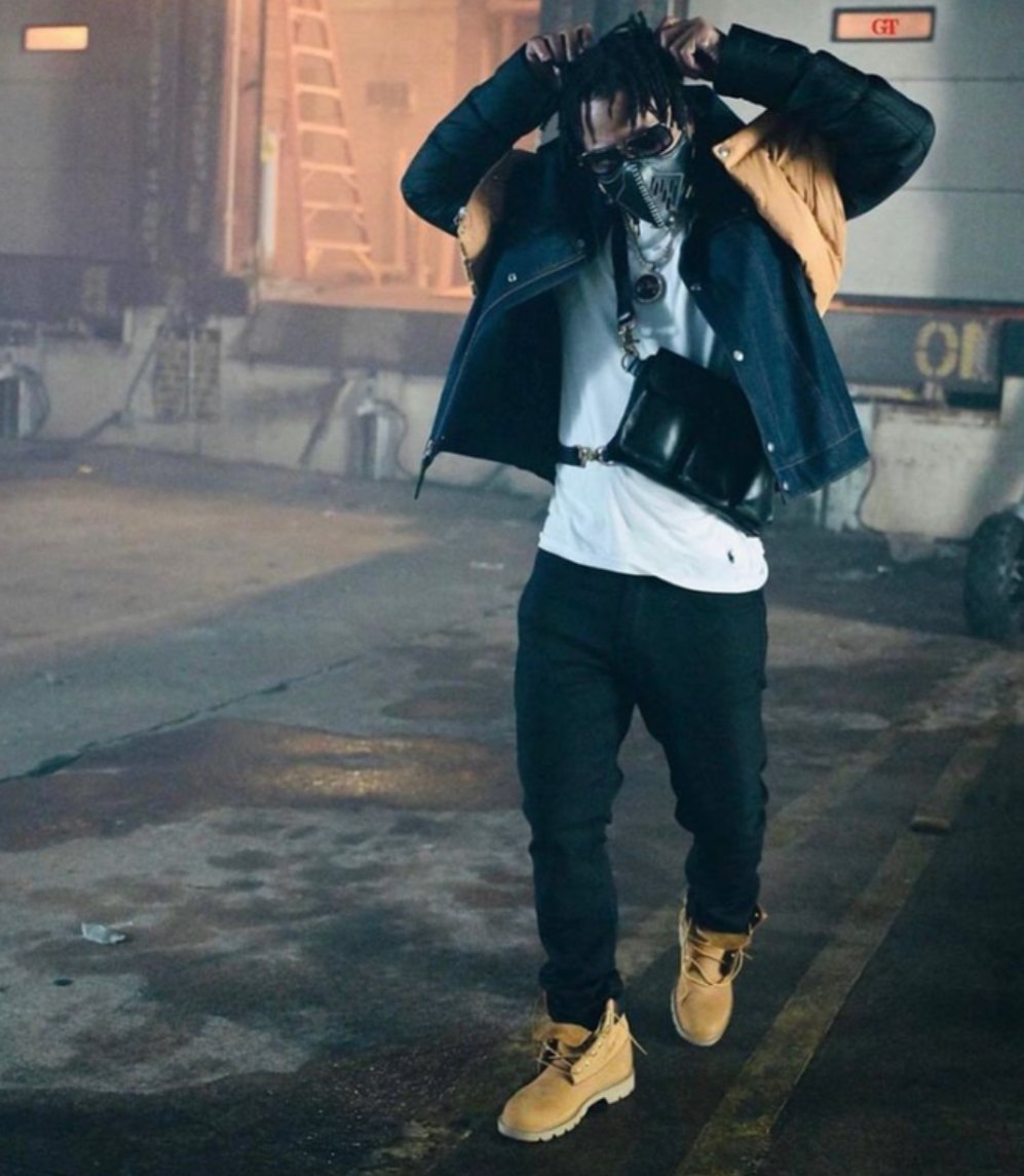 A person with dreadlocks and sunglasses adjusting their hood in a parking lot at night, wearing a black jacket with a brown parka, white T-shirt, black pants, tan work boots, and carrying a black crossbody bag.