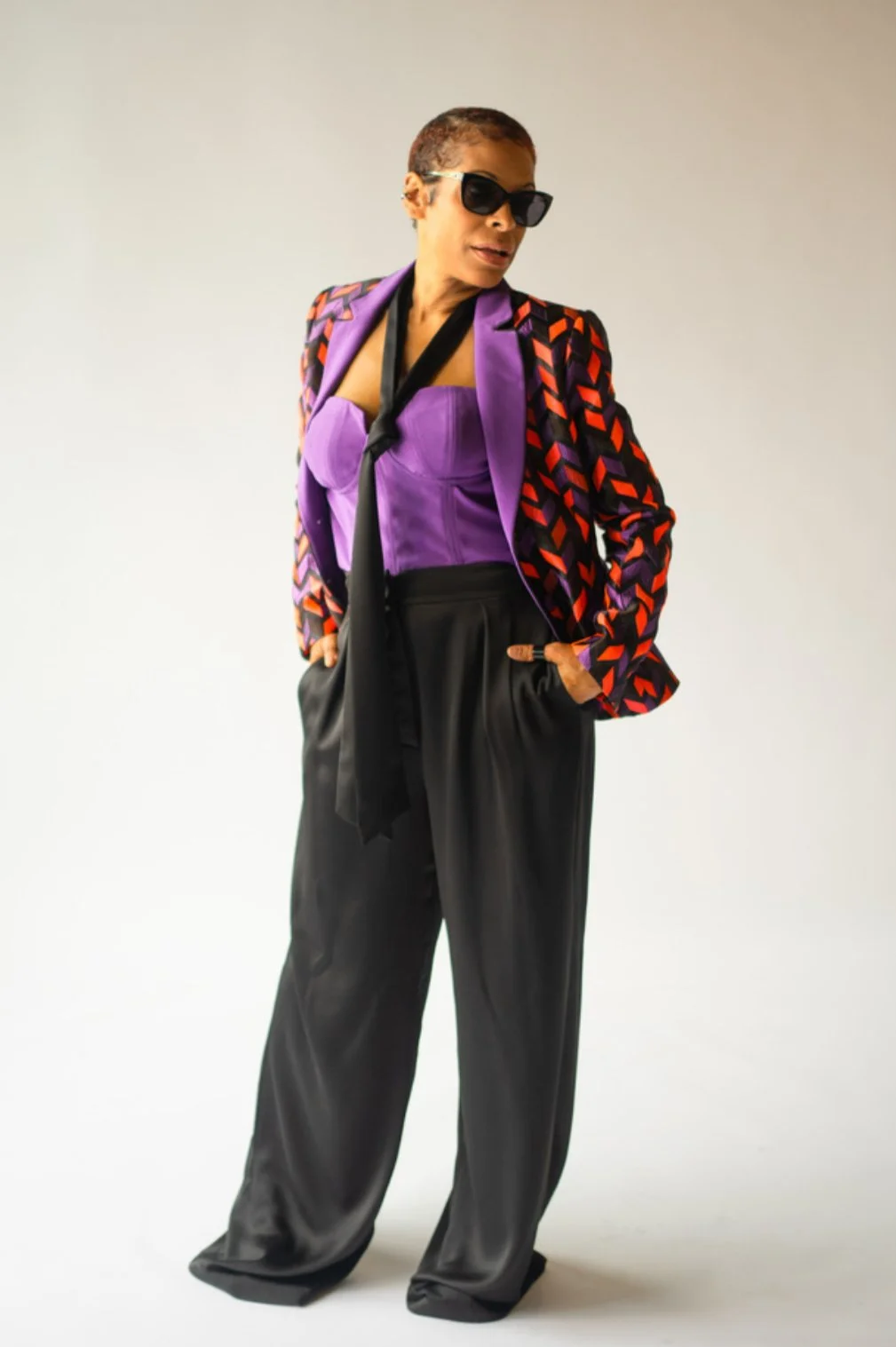 A woman wearing a purple corset top, a patterned blazer, black wide-leg pants, sunglasses, and a black tie around her neck, standing against a plain background.