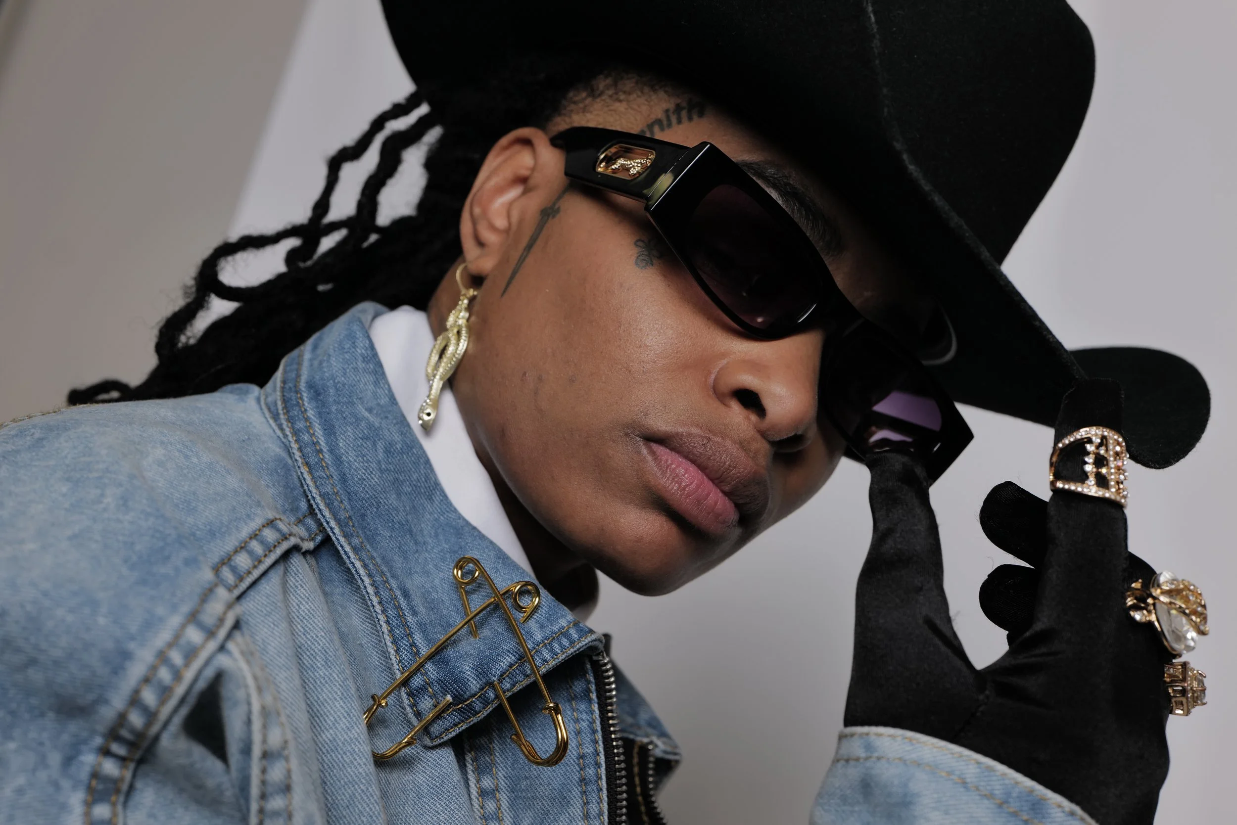 A person wearing a black wide-brimmed hat, dark sunglasses, gold jewelry, denim jacket with safety pins, black gloves, and holding the brim of the hat.
