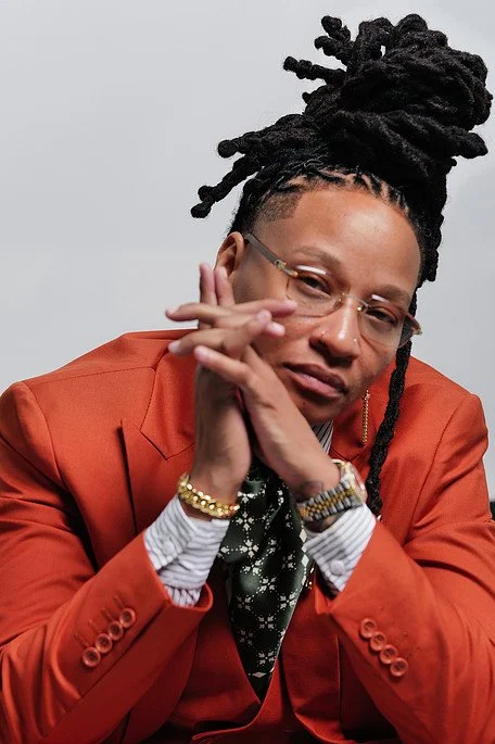 A woman with dreadlocks styled in an updo, wearing glasses, a coral blazer, and white shirt with a black patterned tie, resting her chin on her folded hands, looking at the camera.