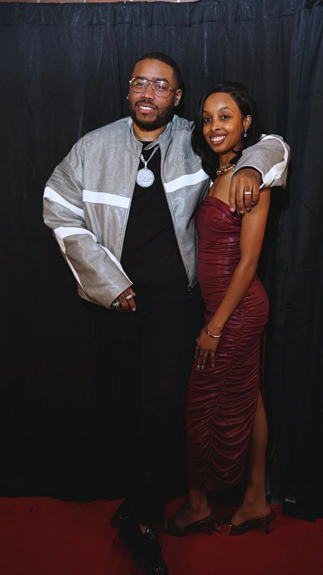 A man and woman posing together in front of a black backdrop. The man is wearing glasses, a silver jacket, and a large pendant necklace. The woman is dressed in a strapless burgundy dress and heels. They are smiling and appear to be at a formal event or party.
