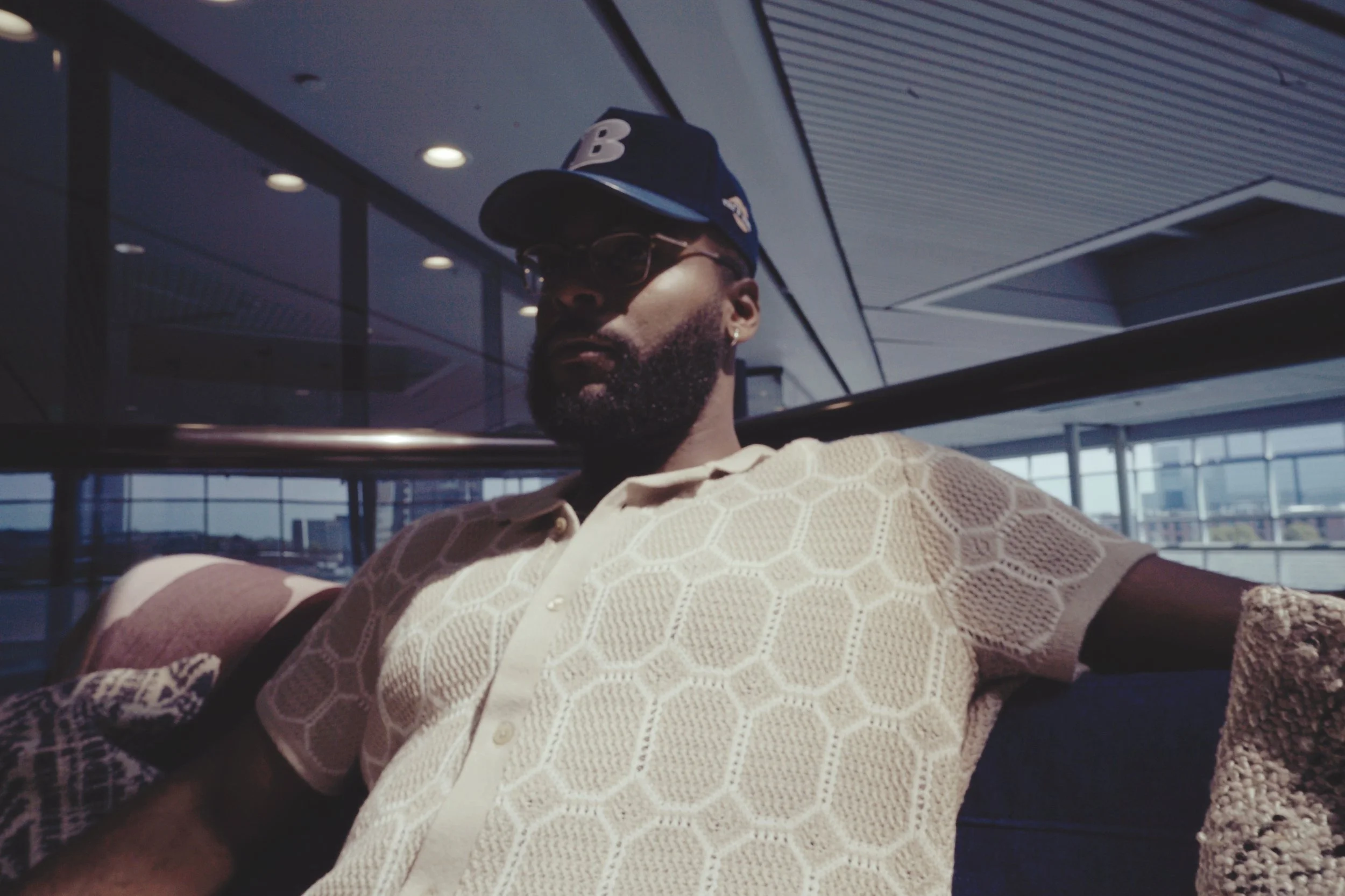 A man with a beard wearing sunglasses, a blue baseball cap, and a cream-colored patterned shirt, sitting on a sofa in an airport waiting area with large windows and cityscape in the background.