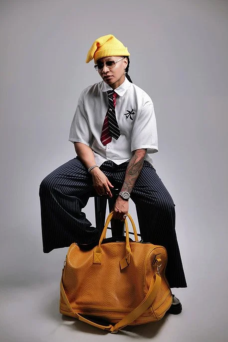 A person sitting on a stool with a yellow handbag in front. They are wearing a yellow beanie, sunglasses, a white striped shirt with a black and red striped tie, dark pinstripe pants, and black shoes.