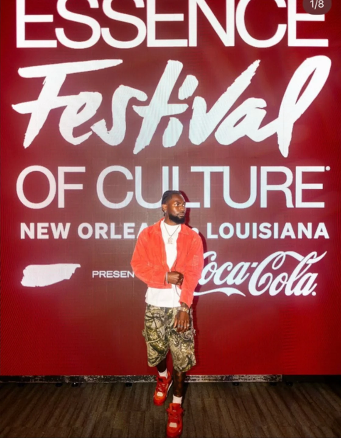 A man standing in front of a red backdrop with white text reading 'Essence Festival of Culture, New Orleans Louisiana' and featuring the Coca-Cola logo. The man is wearing a red jacket, patterned shorts, red sneakers, and jewelry.
