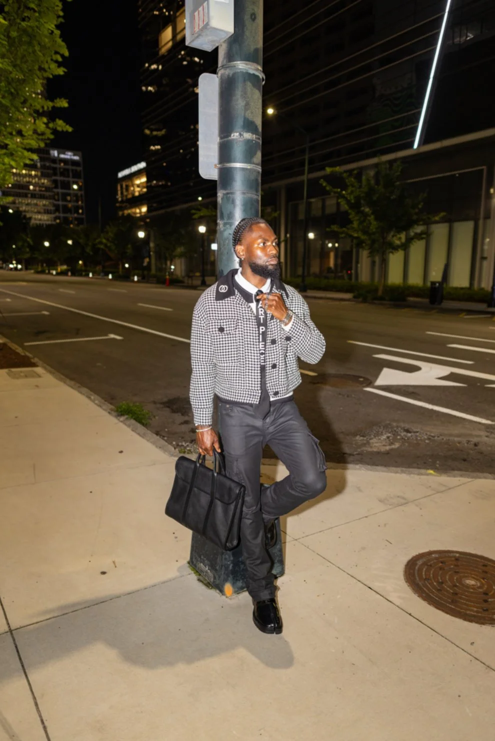 A man standing on a city sidewalk at night, leaning against a streetlamp, holding a black briefcase, wearing a checkered jacket, black cargo pants, and black shoes.