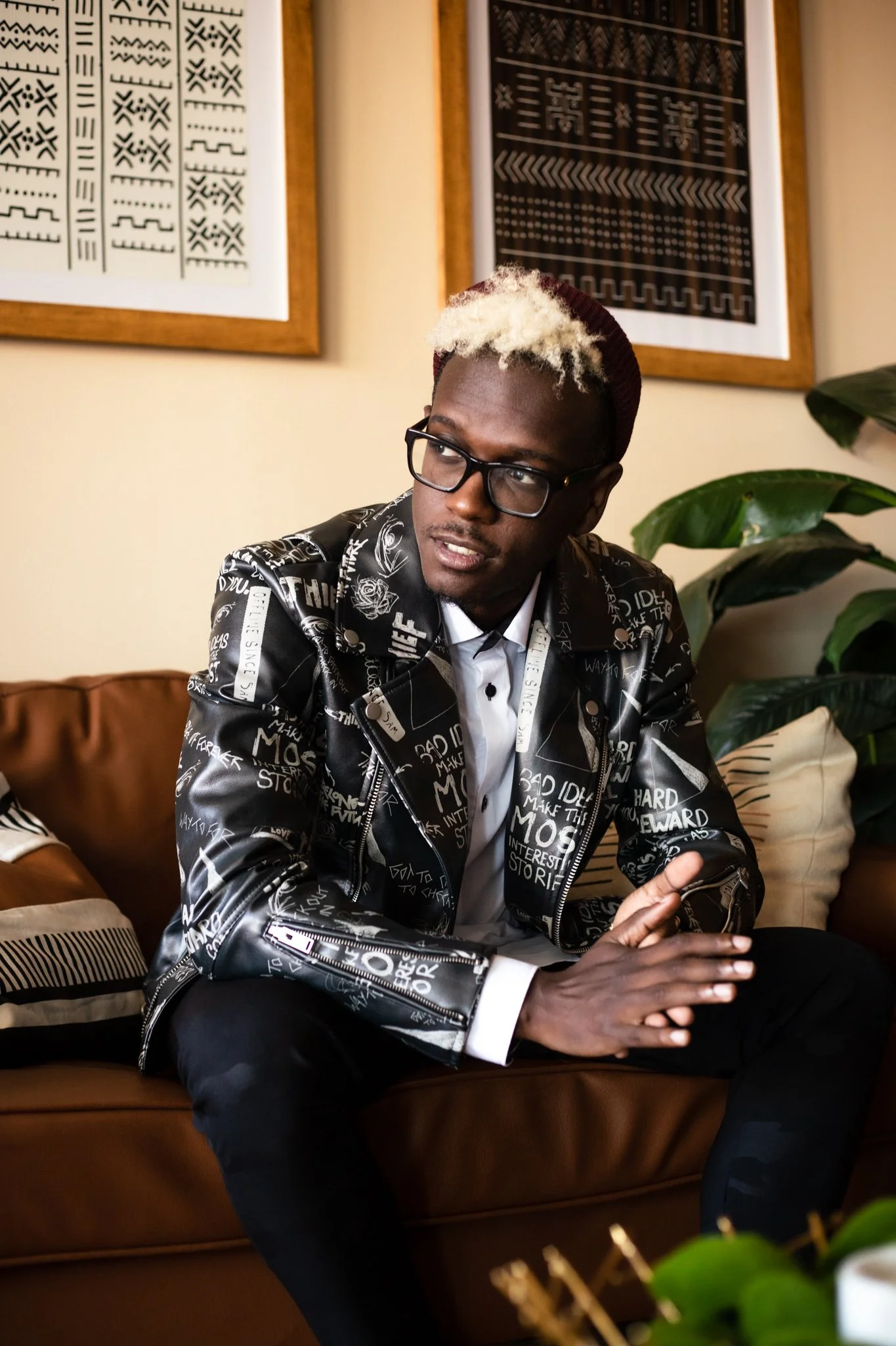 A young man with platinum blonde hair, wearing glasses, a stylish black jacket with white text and symbols, sitting on a brown sofa, surrounded by framed African textile art and green plants.