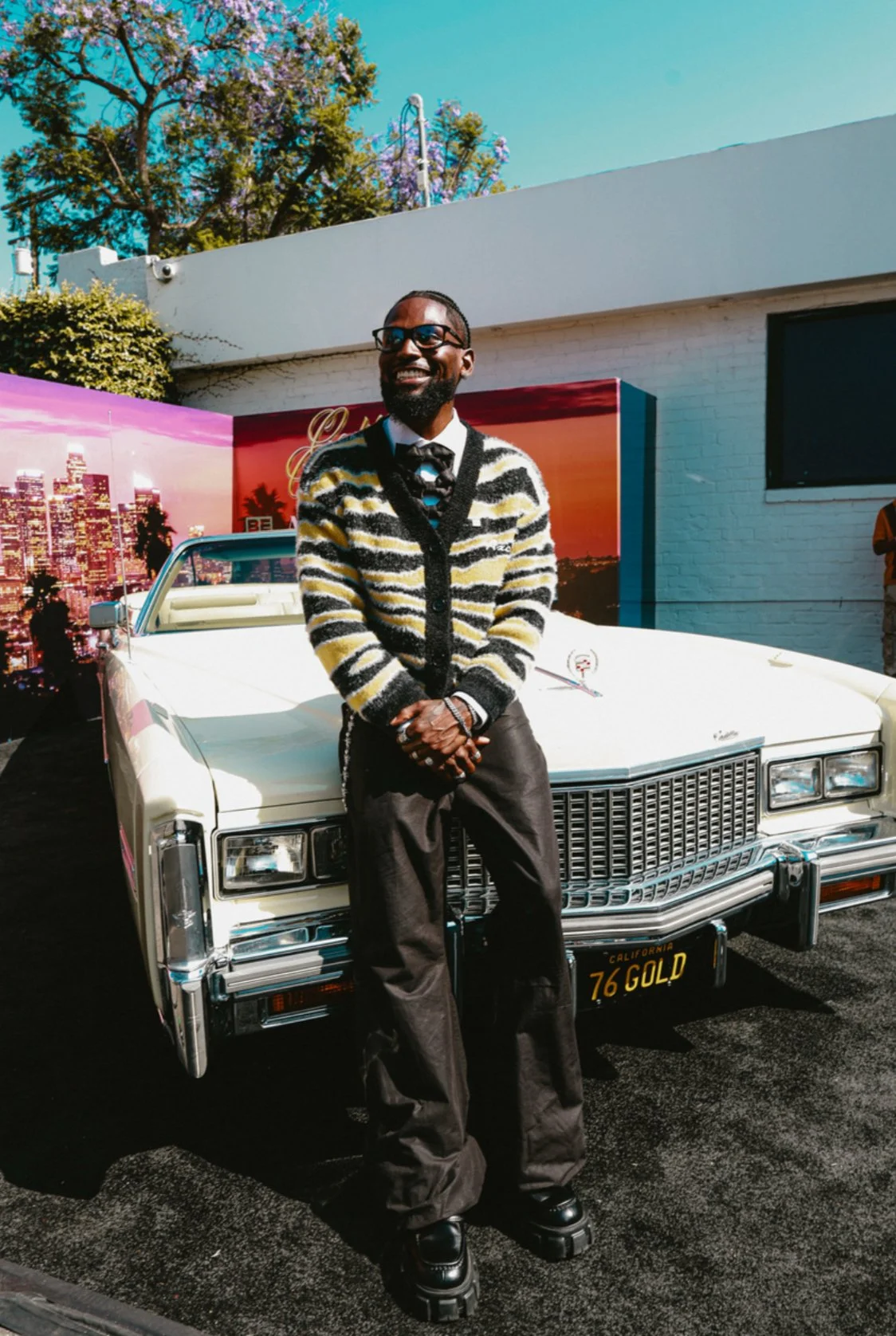 A man standing in front of a vintage white car with a desert sunset mural behind him. He is dressed in a black and yellow striped cardigan, black pants, and black shoes, and is smiling.