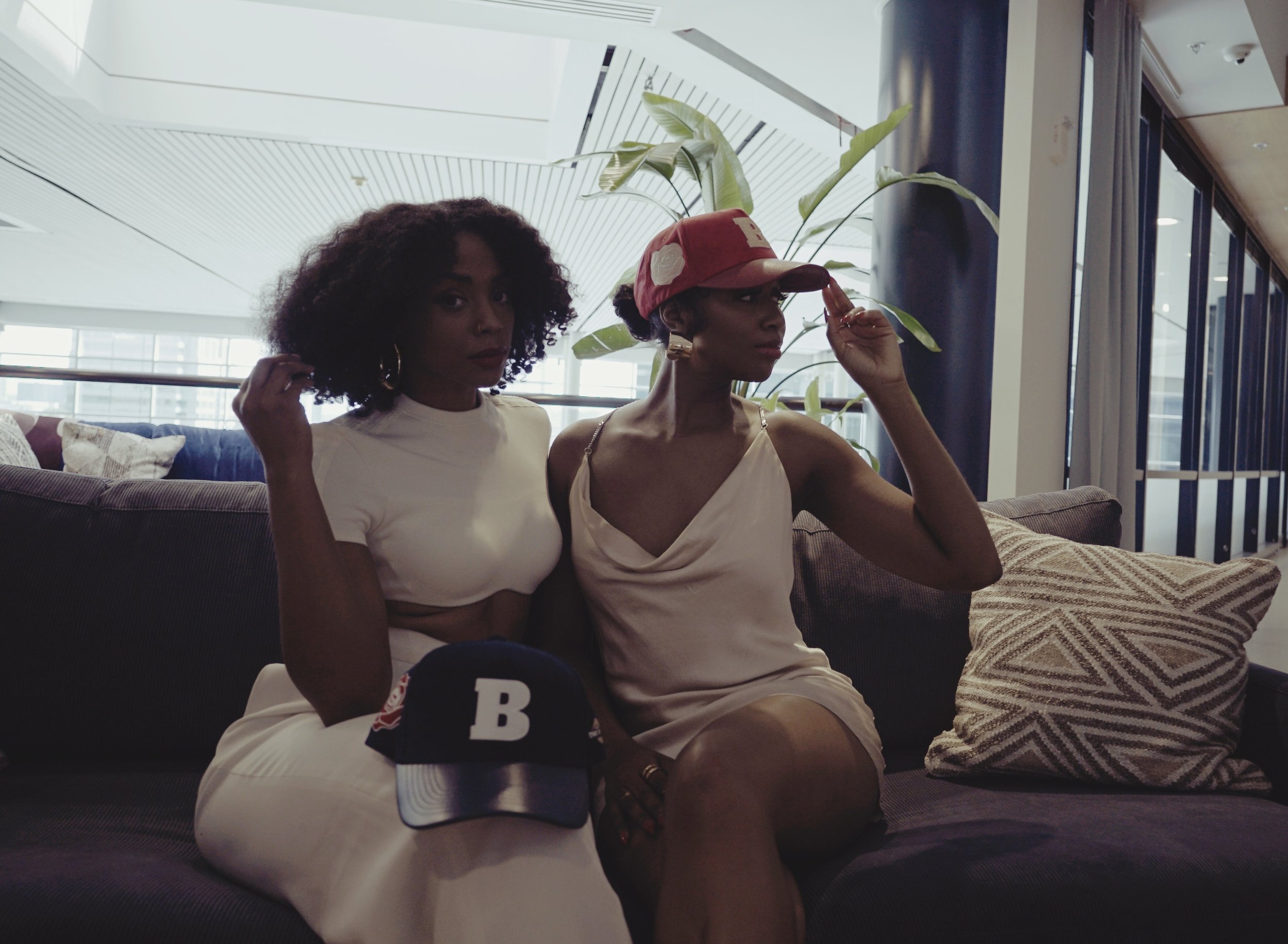 Two women sitting on a dark-colored couch in a modern indoor space, one wearing a white top and the other wearing a light-colored dress, with a baseball cap on her lap and another worn, and a large leafy plant behind them.