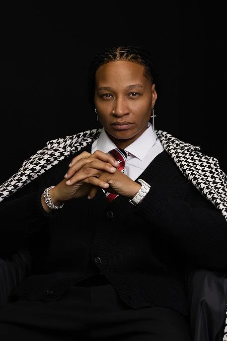 A woman dressed in a black suit with a white collared shirt and red striped tie, sitting with hands clasped, wearing a houndstooth patterned scarf over her shoulders, and sporting jewelry including earrings, a watch, and a bracelet, against a black b