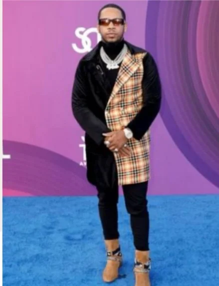 Man wearing sunglasses, black jacket with a plaid pattern, dark pants, and tan boots, standing on a blue carpet in front of a purple backdrop.
