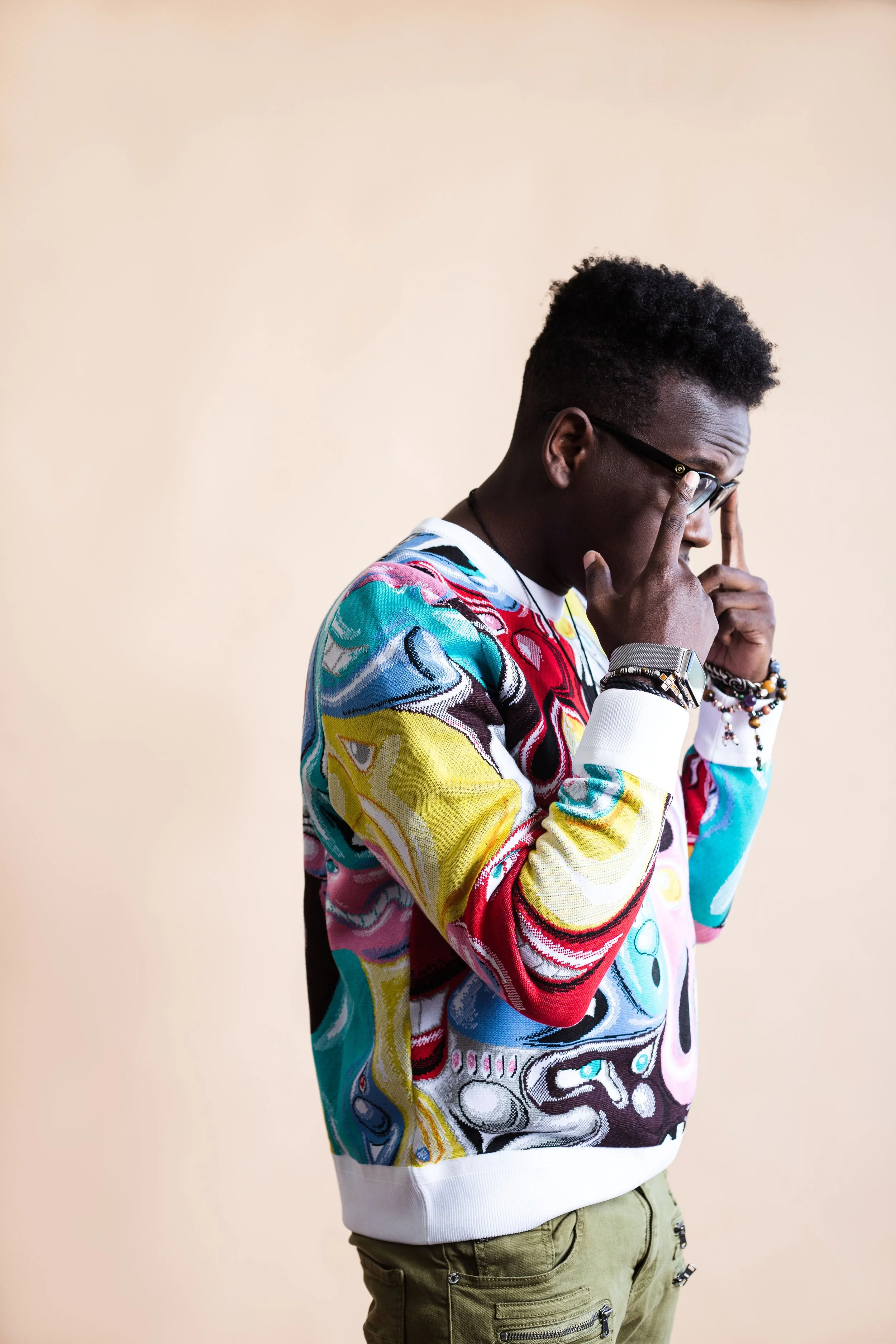 A young man with dark skin and short curly black hair wearing glasses, a colorful patterned sweater, and olive green pants, holding his glasses and looking contemplative against a plain beige background.