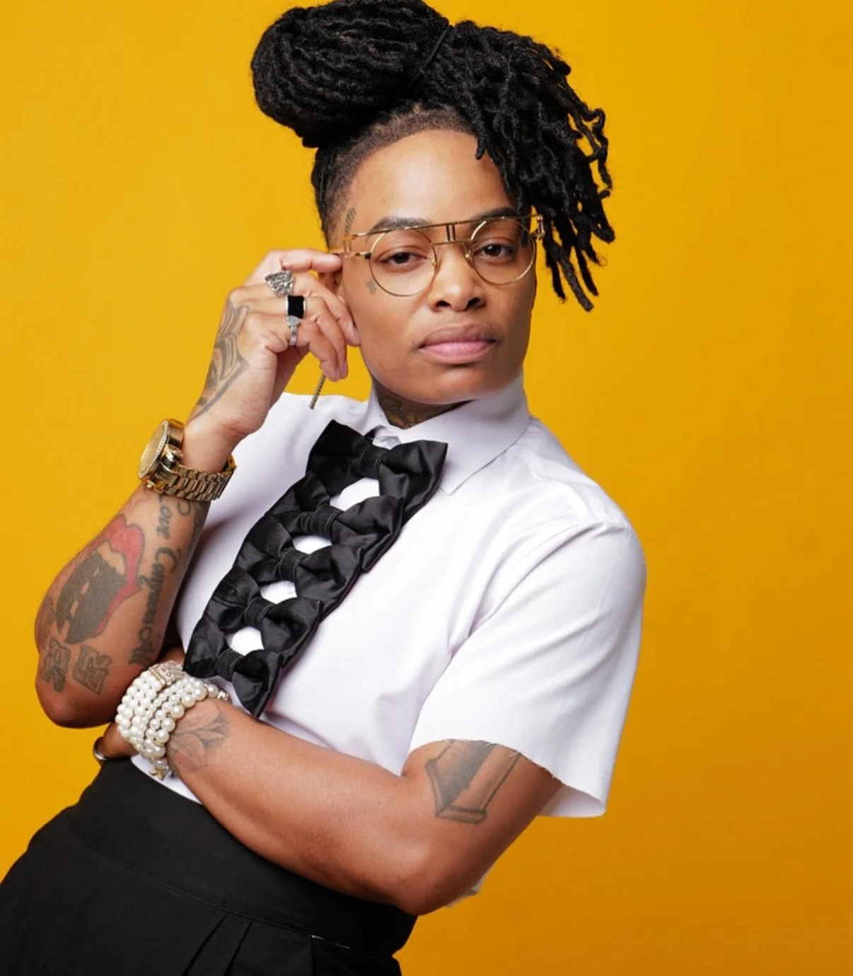 Person with black dreadlocks styled up, wearing glasses, a white shirt with black bow tie, jewelry, and tattoos, against a yellow background.