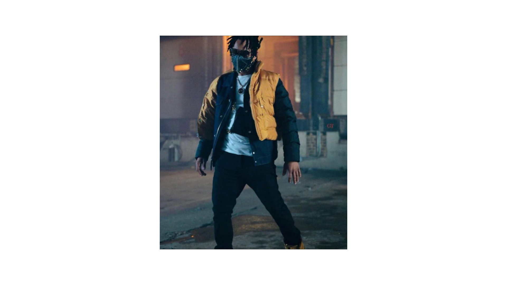 A man wearing a yellow and black puffer jacket, black pants, and yellow shoes, standing outdoors at night with a mask and sunglasses on, in an urban environment.