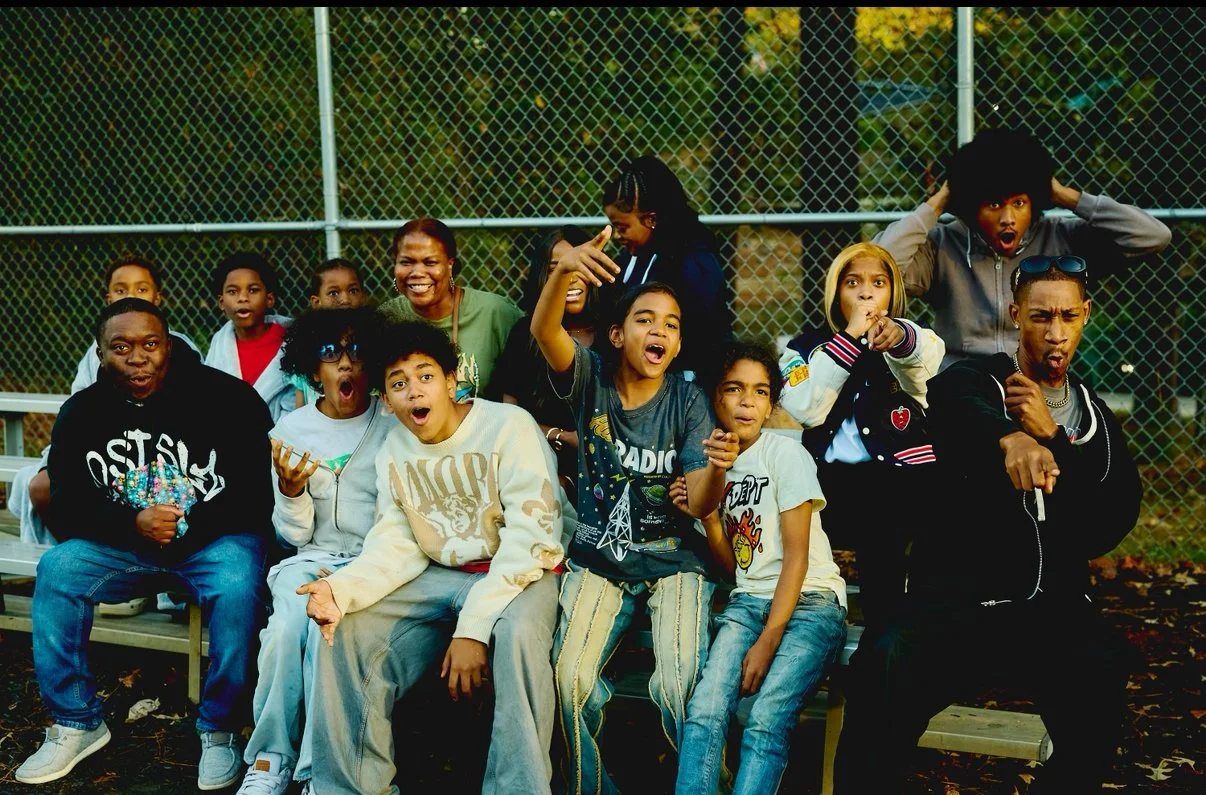 Group of diverse children and adults sitting on a bench at a park, making funny faces and poses.