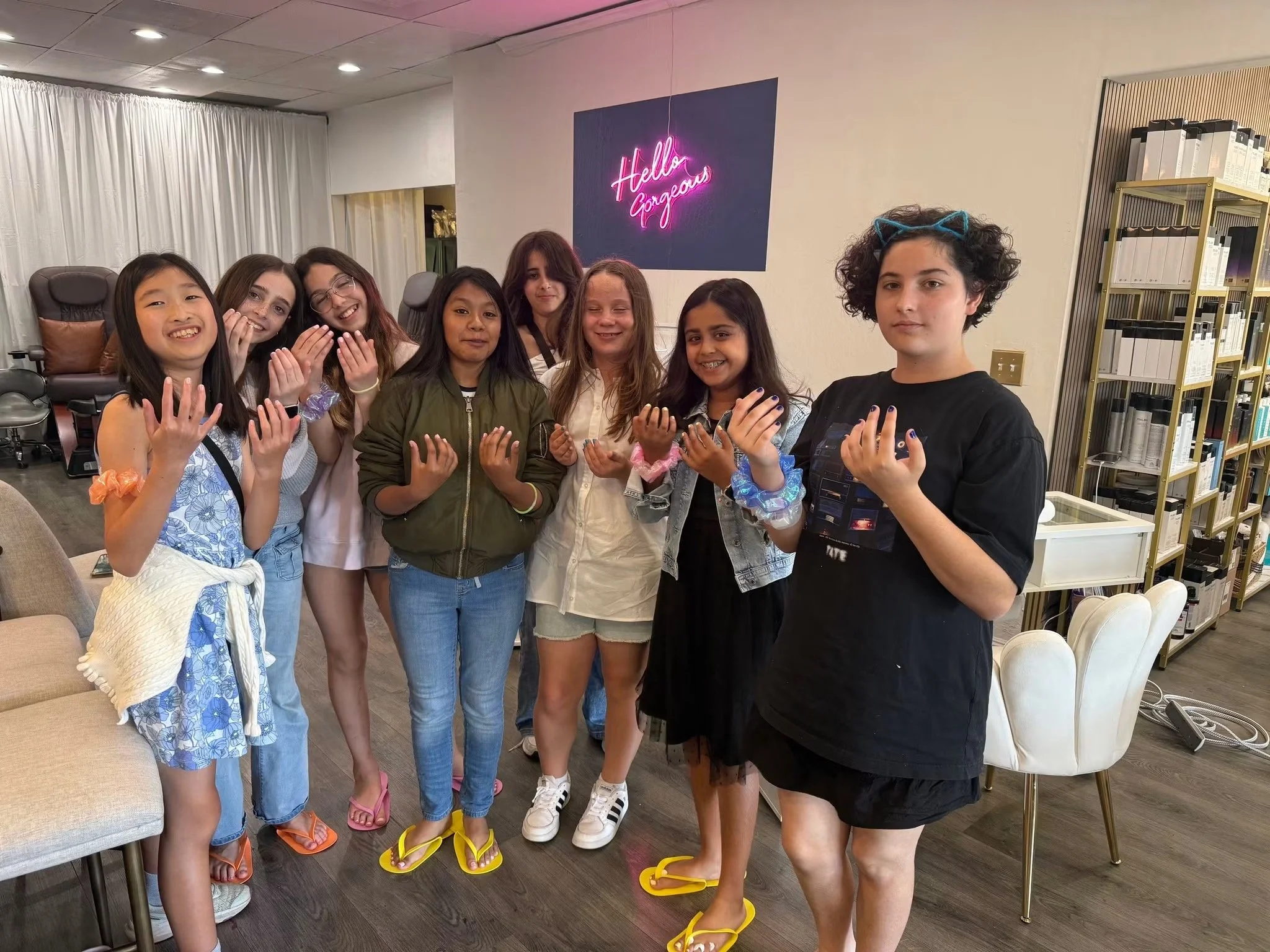Group of girls standing in a salon, smiling and showing their hands after each getting mani pedis and having a party hosted in the salon. A neon sign on the wall behind them says 'Hello Gorgeous'.