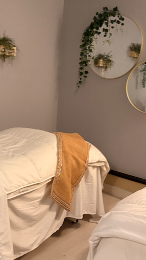 A cozy bedroom corner with two beds, beige blankets, and a mustard-colored throw blanket. Decor includes round mirrors and hanging greenery on the wall.