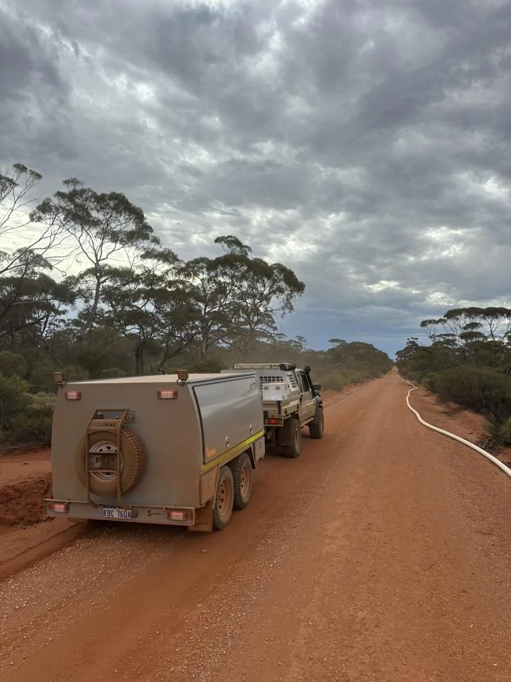 A dirt road in a remote area with an overcast sky, featuring a large utility truck towing a portable water or fuel tank, with sparse trees along the sides.