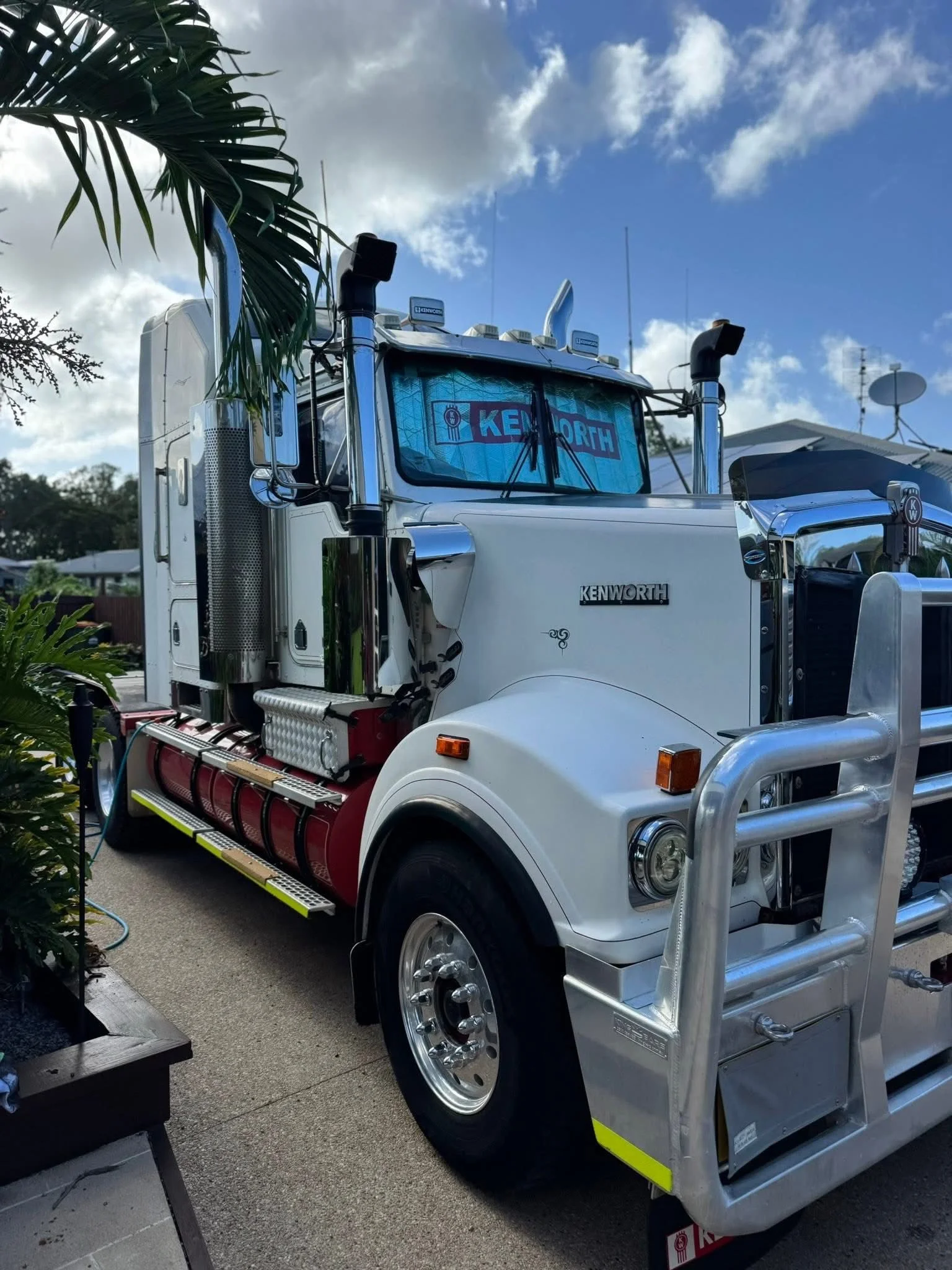 A white Kenworth semi-truck parked outdoors with a blue sky and clouds above. Trees and plants are visible around the truck. The truck features chrome details, including exhaust pipes and a front grille, with the Kenworth logo on the front.