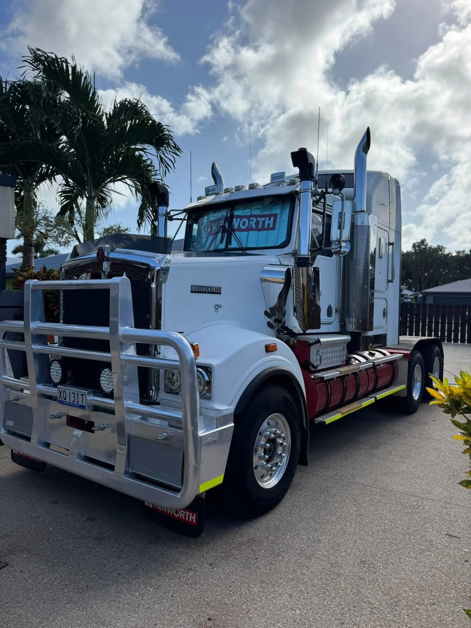 White Kenworth semi-truck with chrome accents parked outdoors under a partly cloudy sky, with a palm tree in the background.