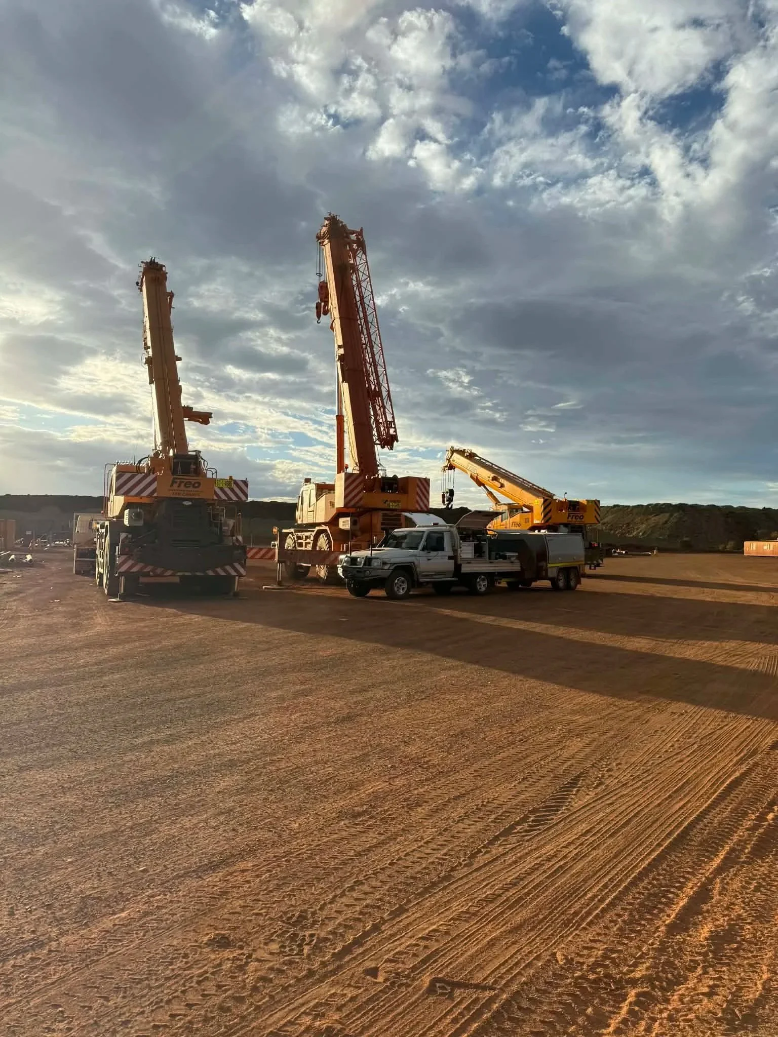 Construction site with three large cranes and a pickup truck on a dirt lot, under a partly cloudy sky during sunset.