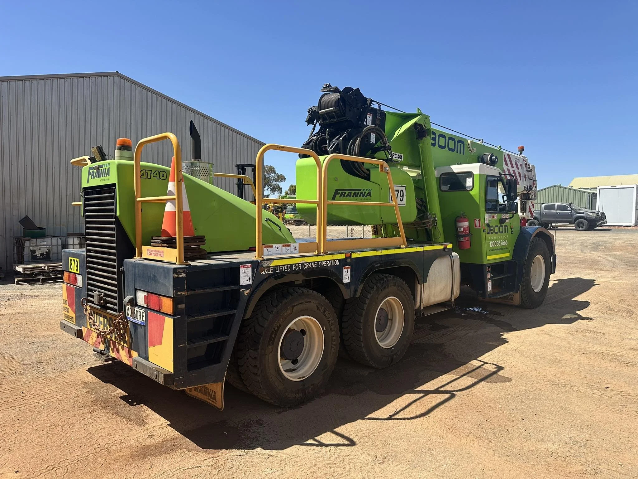 Green crane truck parked on dirt ground with a metal building and vehicles in the background under a clear blue sky.