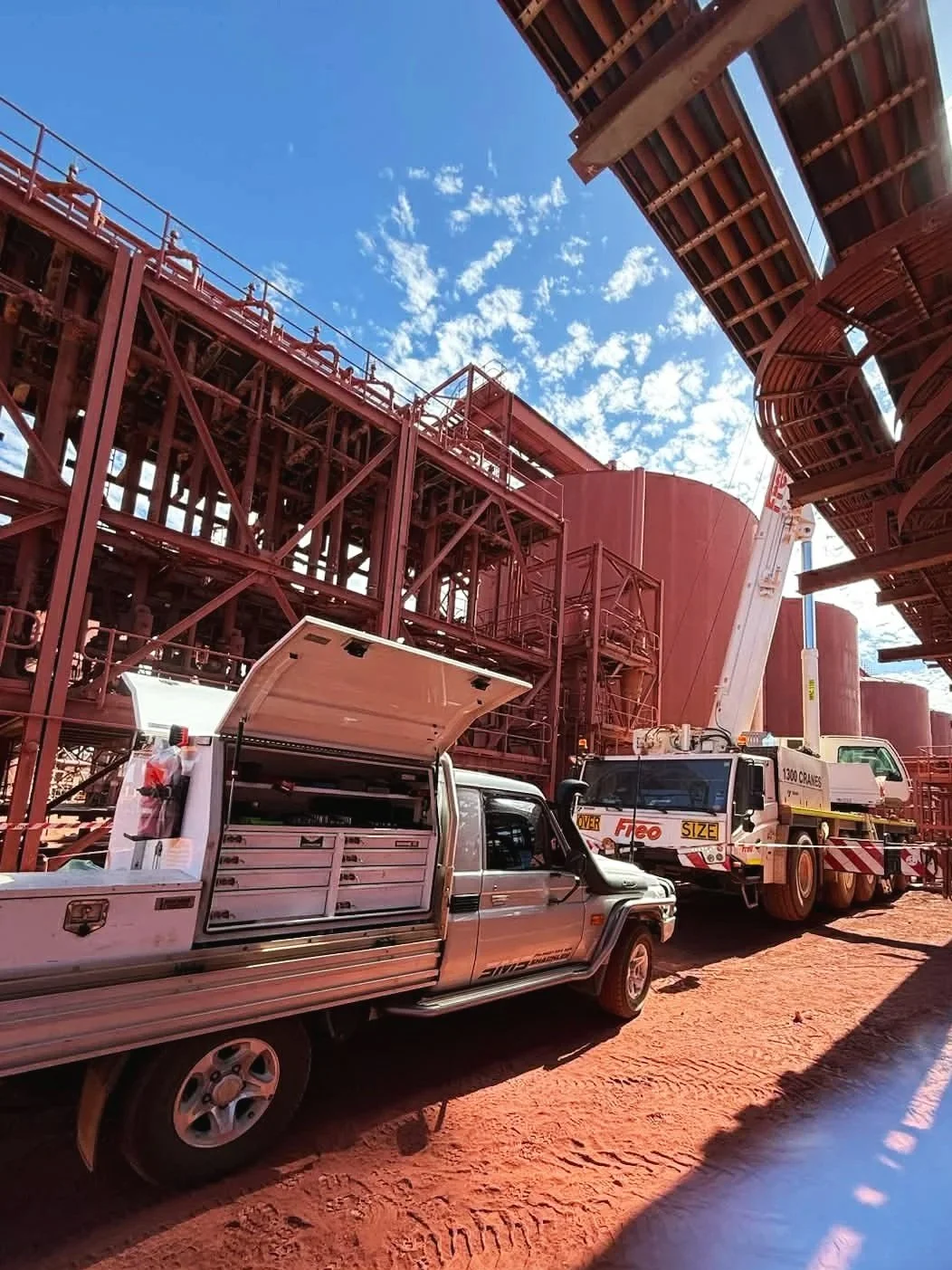 Construction site with red steel structures, a white utility truck with work tools, and a large piece of machinery, under a blue sky with scattered clouds.