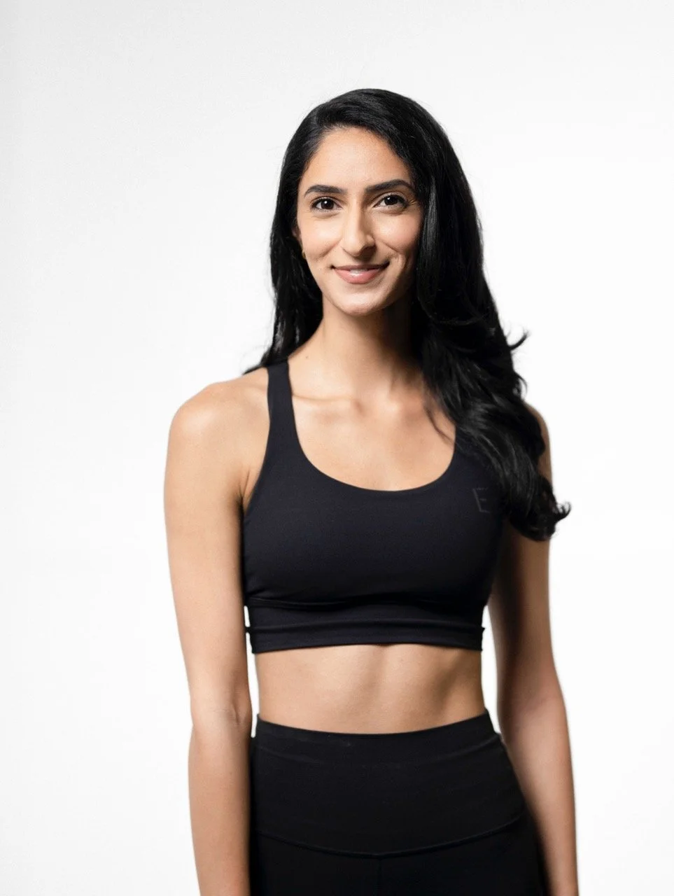 A woman with long black hair wearing a black sports bra and black leggings, standing against a plain white background.
