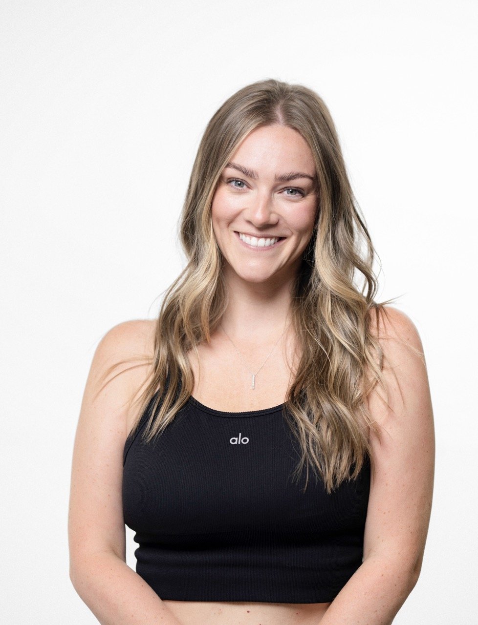 A woman with long wavy blonde hair smiling, wearing a black tank top with 'alo' written on it, standing against a white background.