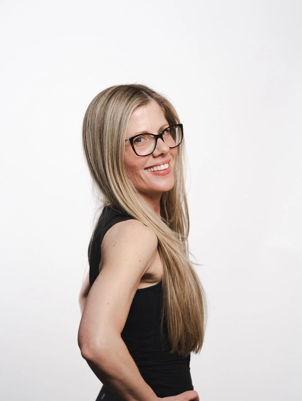 A woman with long blonde hair, wearing glasses and a sleeveless black top, smiling and posing against a plain white background.