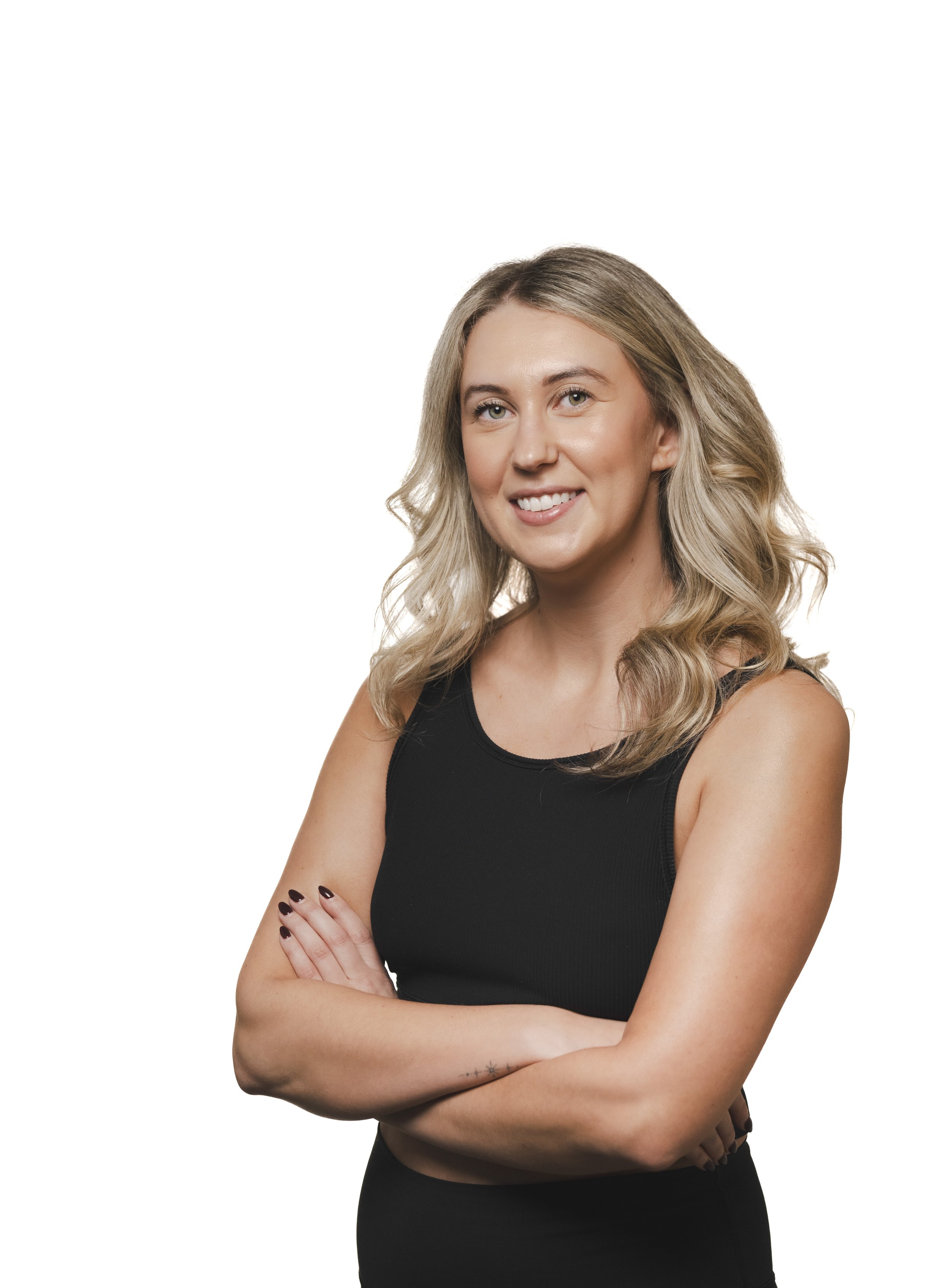 A young woman with blonde, wavy hair wearing a sleeveless black top, smiling with arms crossed, against a plain white background.