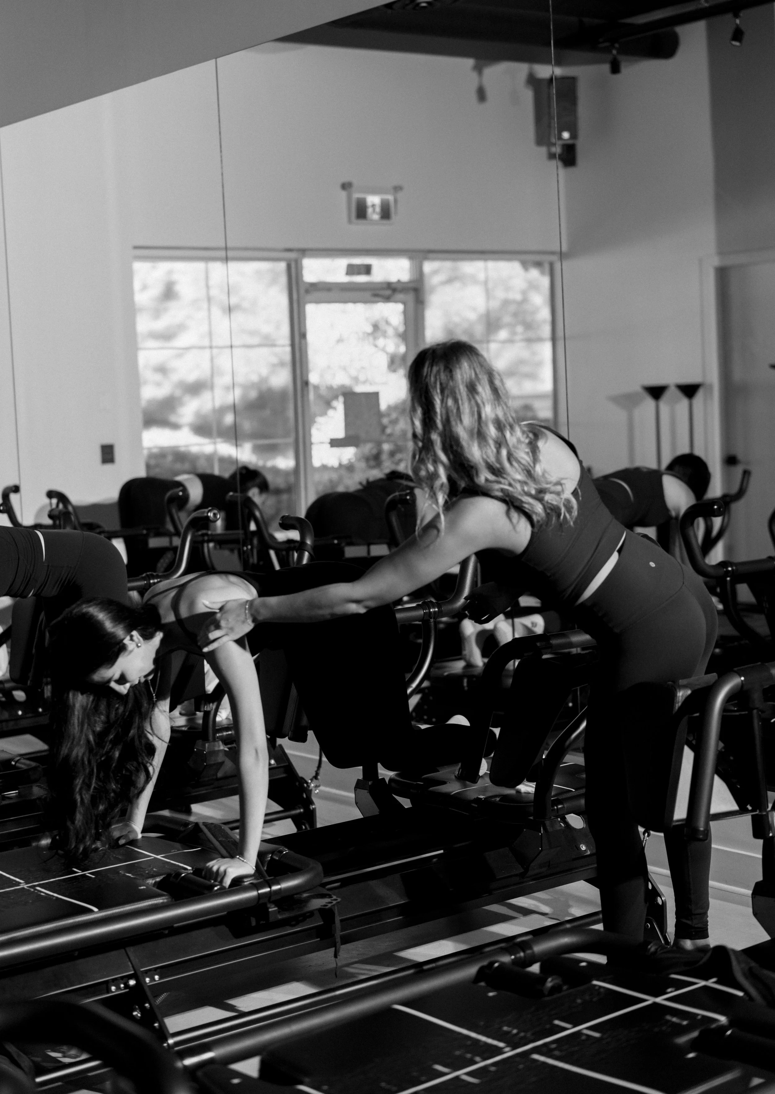 A woman in a fitness class assisting another woman with a workout on a reformer machine inside a gym or fitness studio.