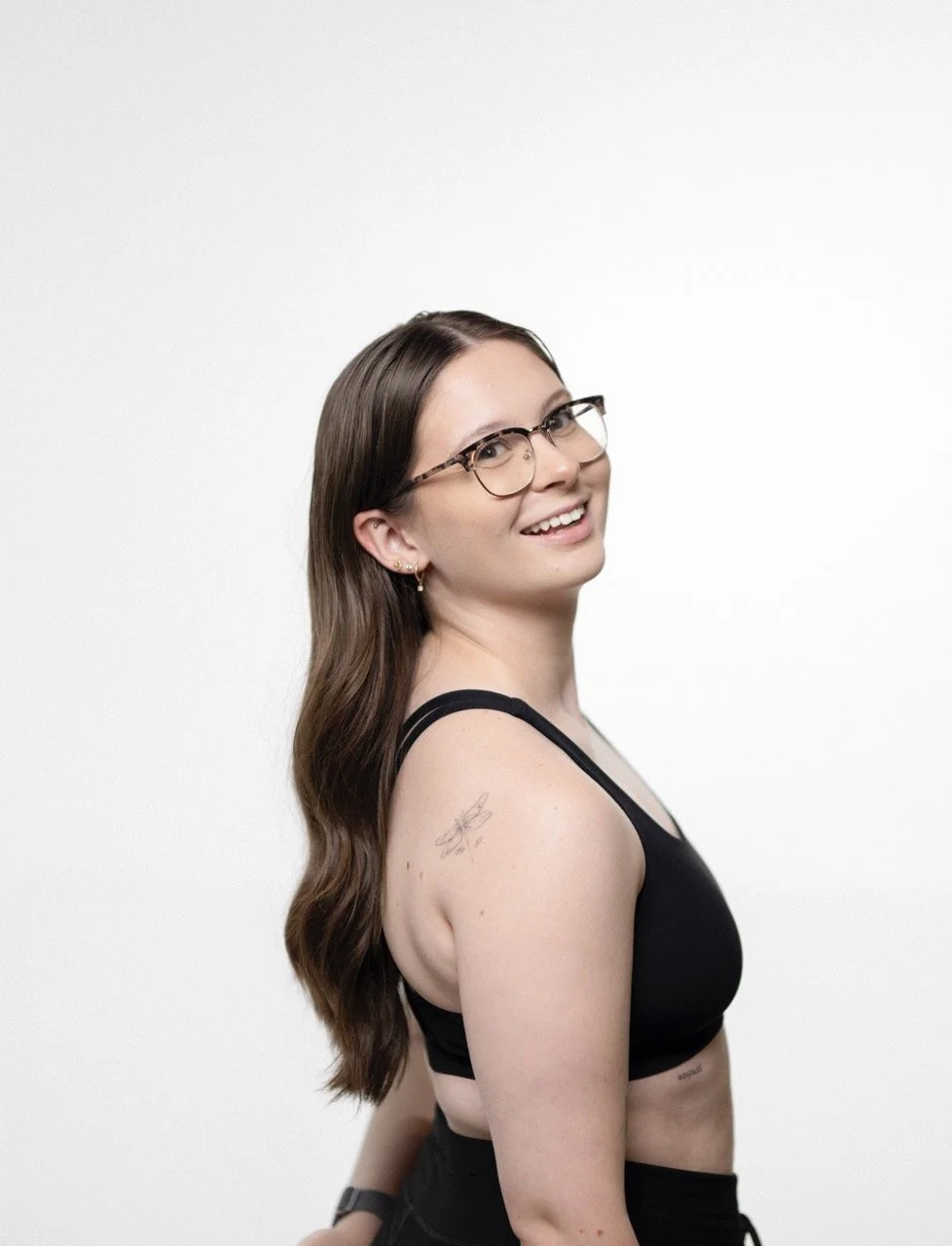 Smiling woman with long brown hair, glasses, a black sports bra, and a tattoo on her left shoulder, standing against a white background.