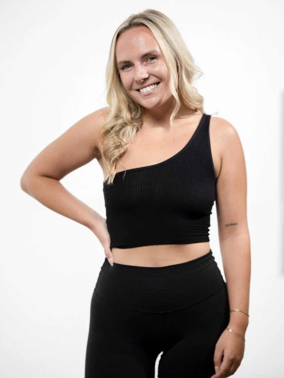 A smiling woman with blonde hair wearing a black tank top and black pants, standing against a plain white background.