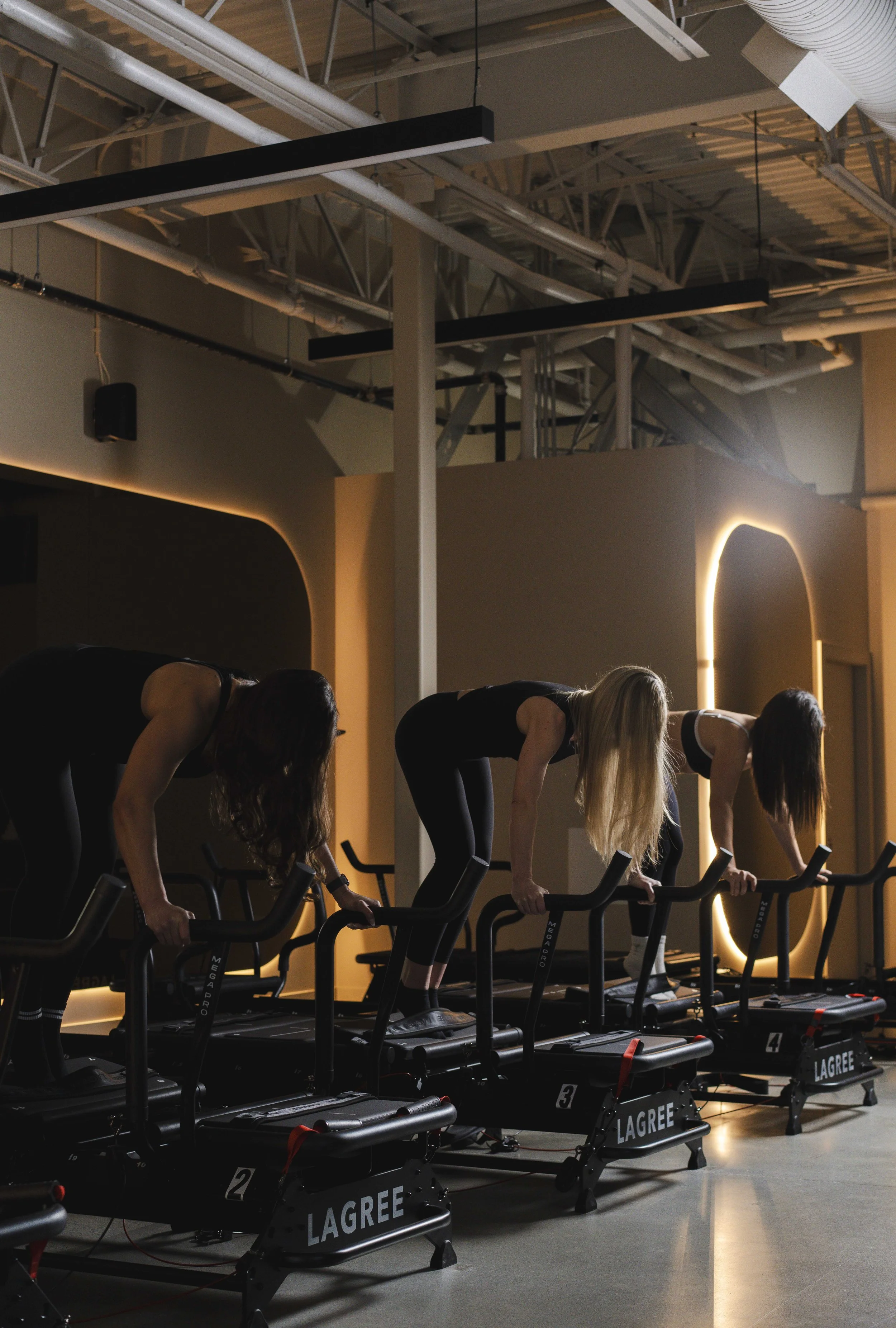 Three women in athletic clothing using rowing machines in a modern gym with lighting and exposed ceiling ductwork.