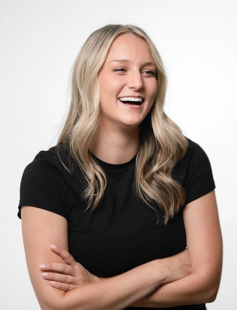 A woman with long blonde hair, wearing a black t-shirt, laughing with her arms crossed against a plain white background.