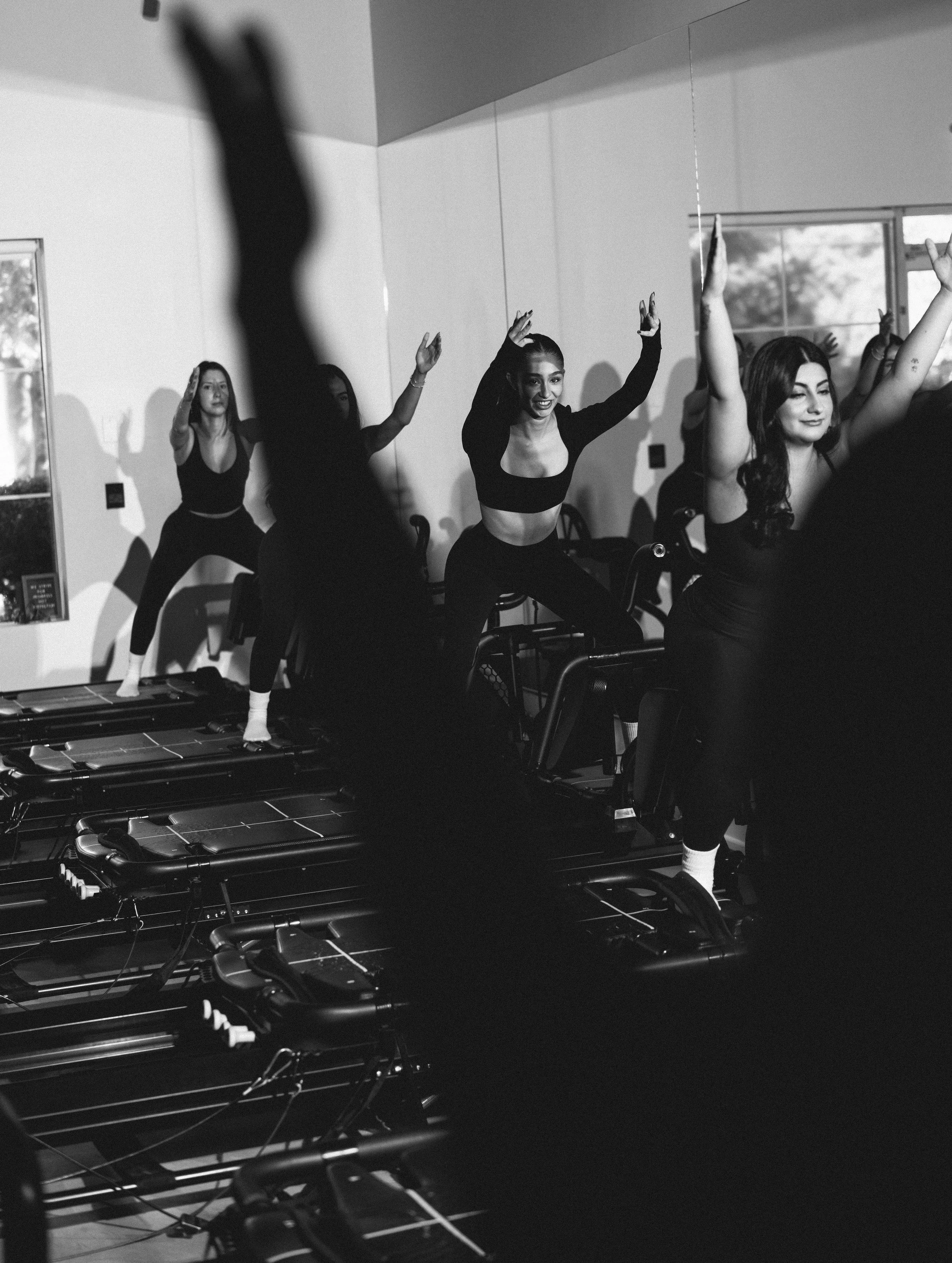 Women participating in a lively indoor fitness class on exercise machines, with some raising their arms and smiling.