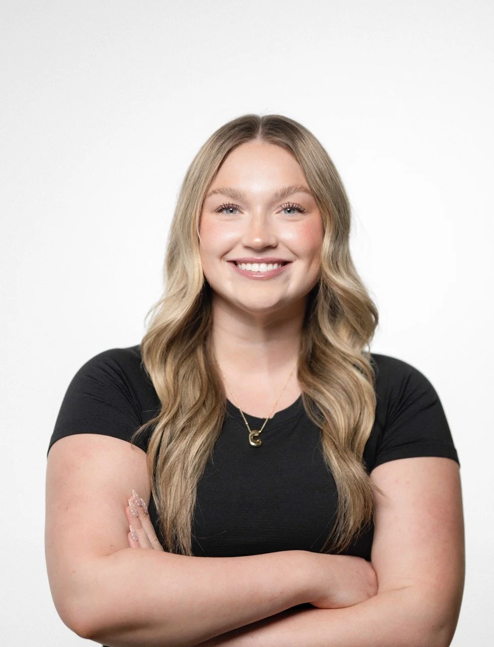 Young woman with long blonde hair smiling, arms crossed, wearing a black t-shirt and a gold necklace, against a plain white background.
