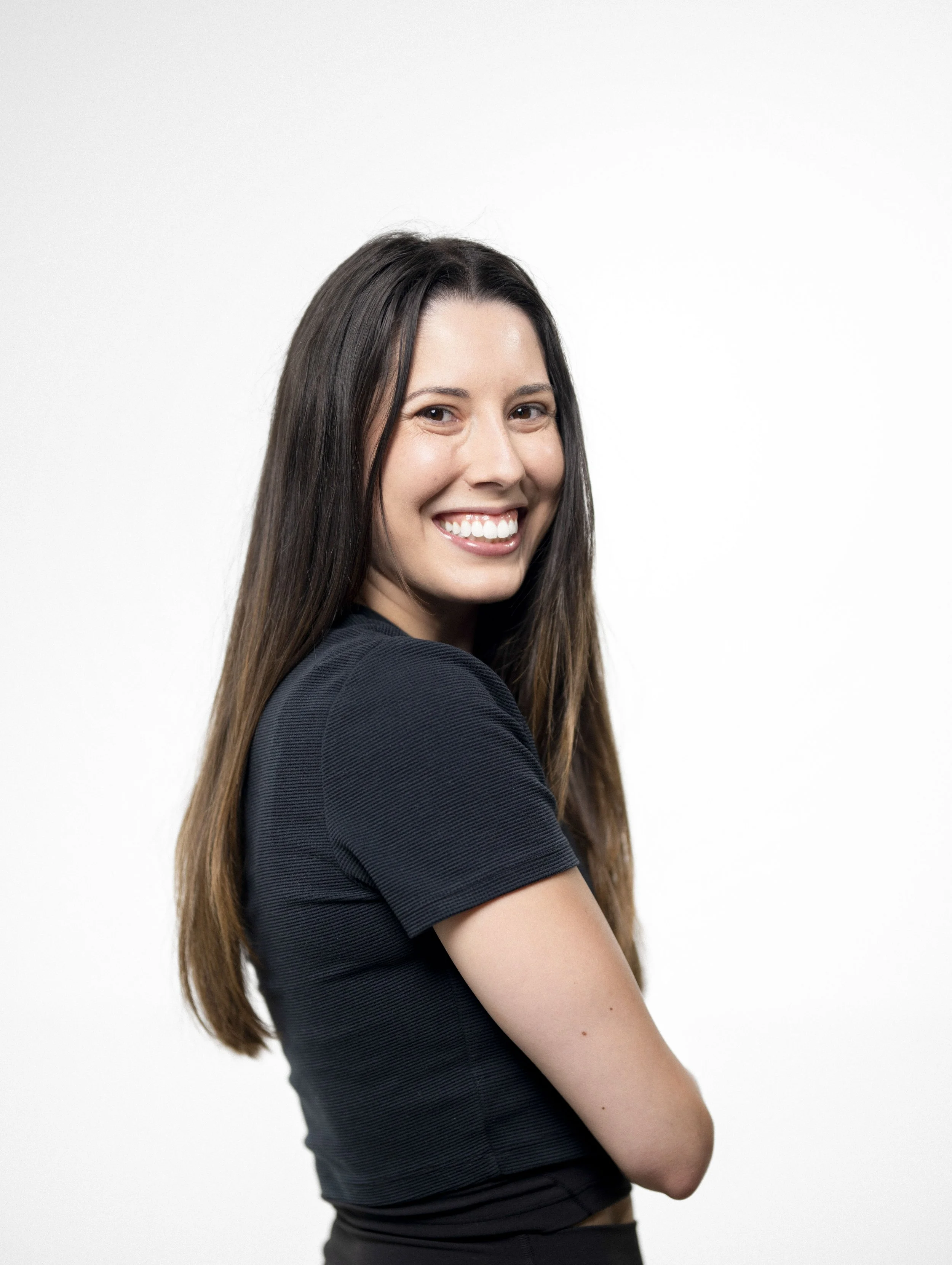 A young woman with long brown hair smiling and looking at the camera, wearing a black shirt, against a plain white background.