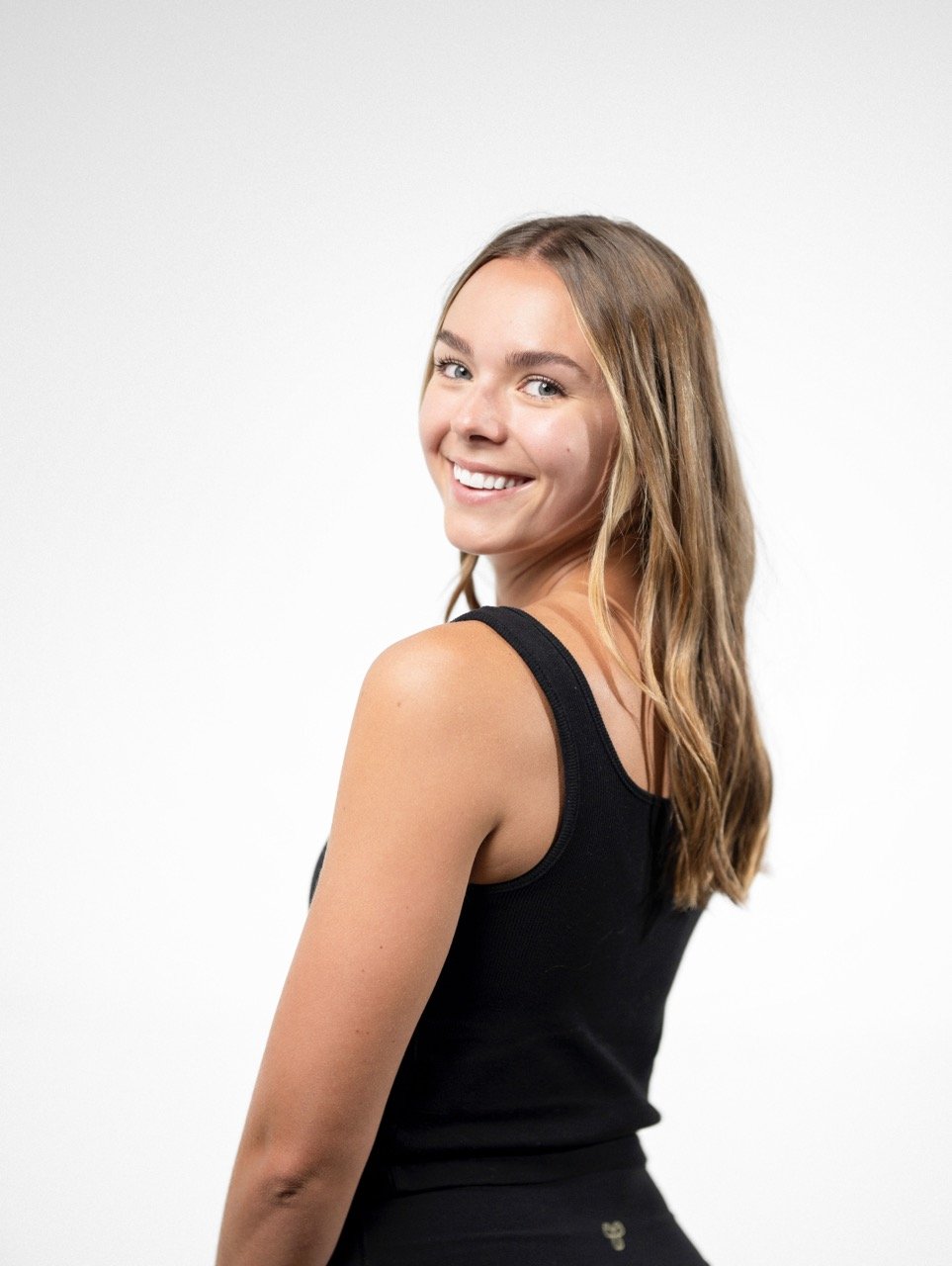 Young woman with long wavy hair smiling at camera, wearing a black sleeveless top, standing against a plain white background.