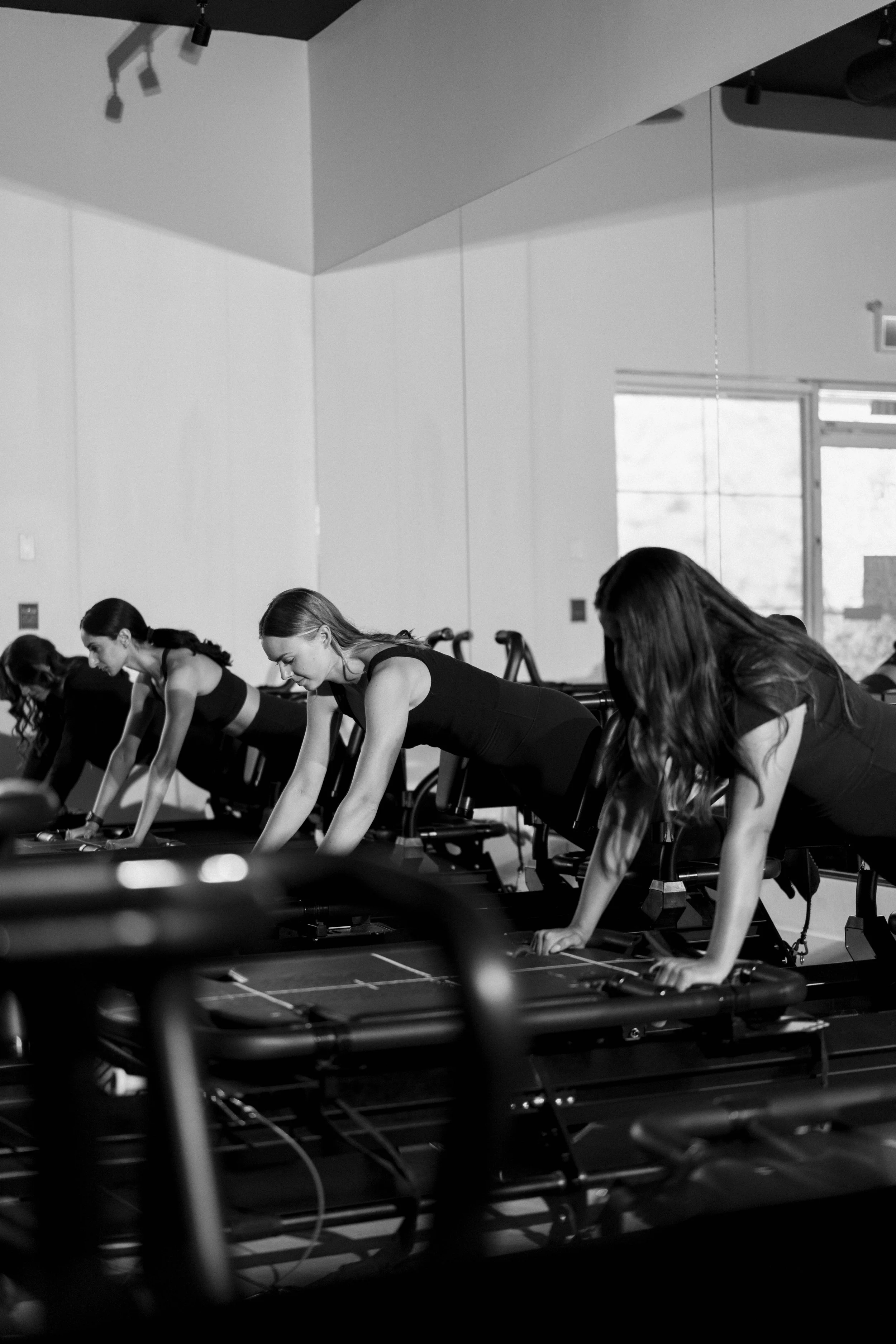 Four women in a gym participating in a pilates class, performing exercises on reformer machines, in a black and white photo.