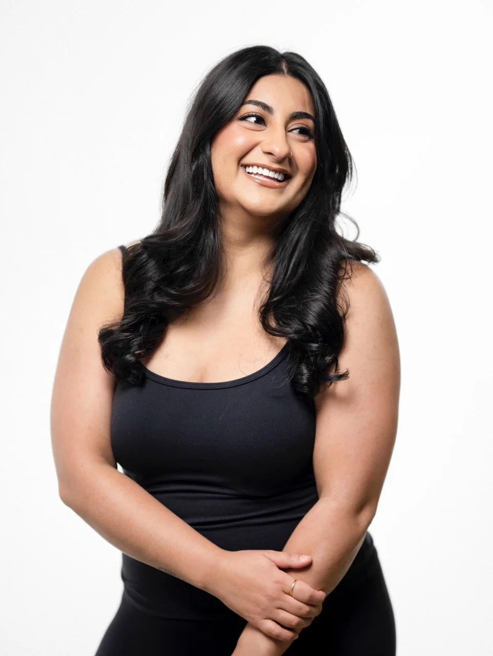 A smiling woman with long black hair wearing a black sleeveless top and standing against a white background.