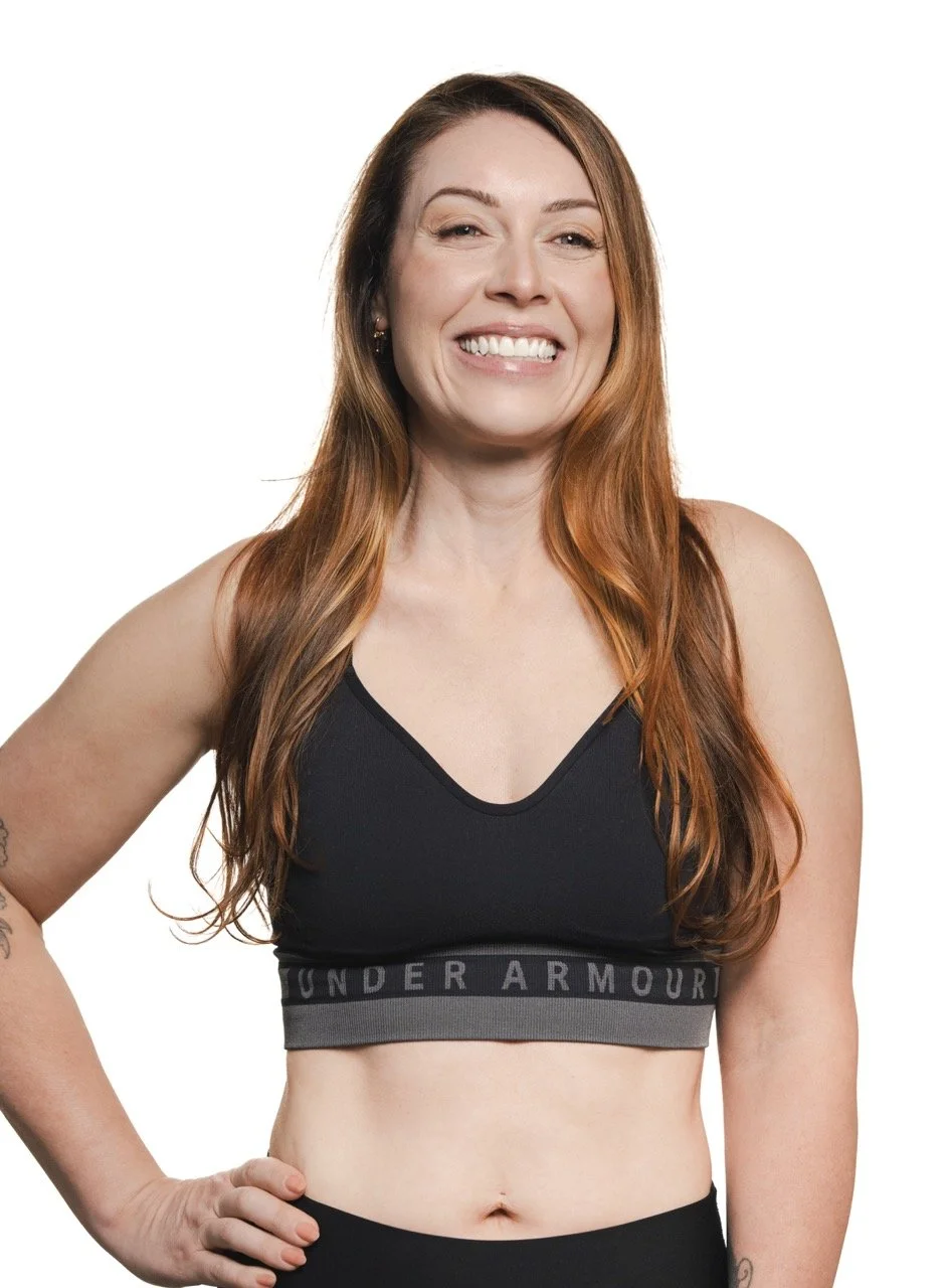 A smiling woman with long red hair wearing a black sports bra with 'Under Armour' branding and black pants, posing with her right hand on her hip against a white background.