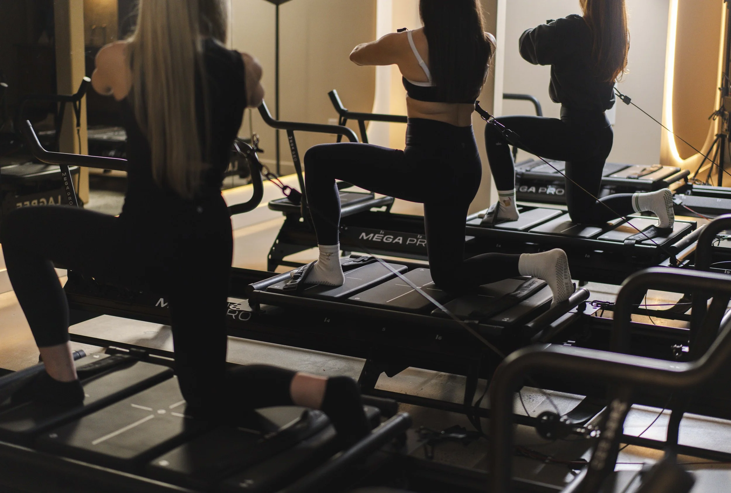 Three women are participating in a Pilates reformer workout class in a gym, stretching on Pilates machines.