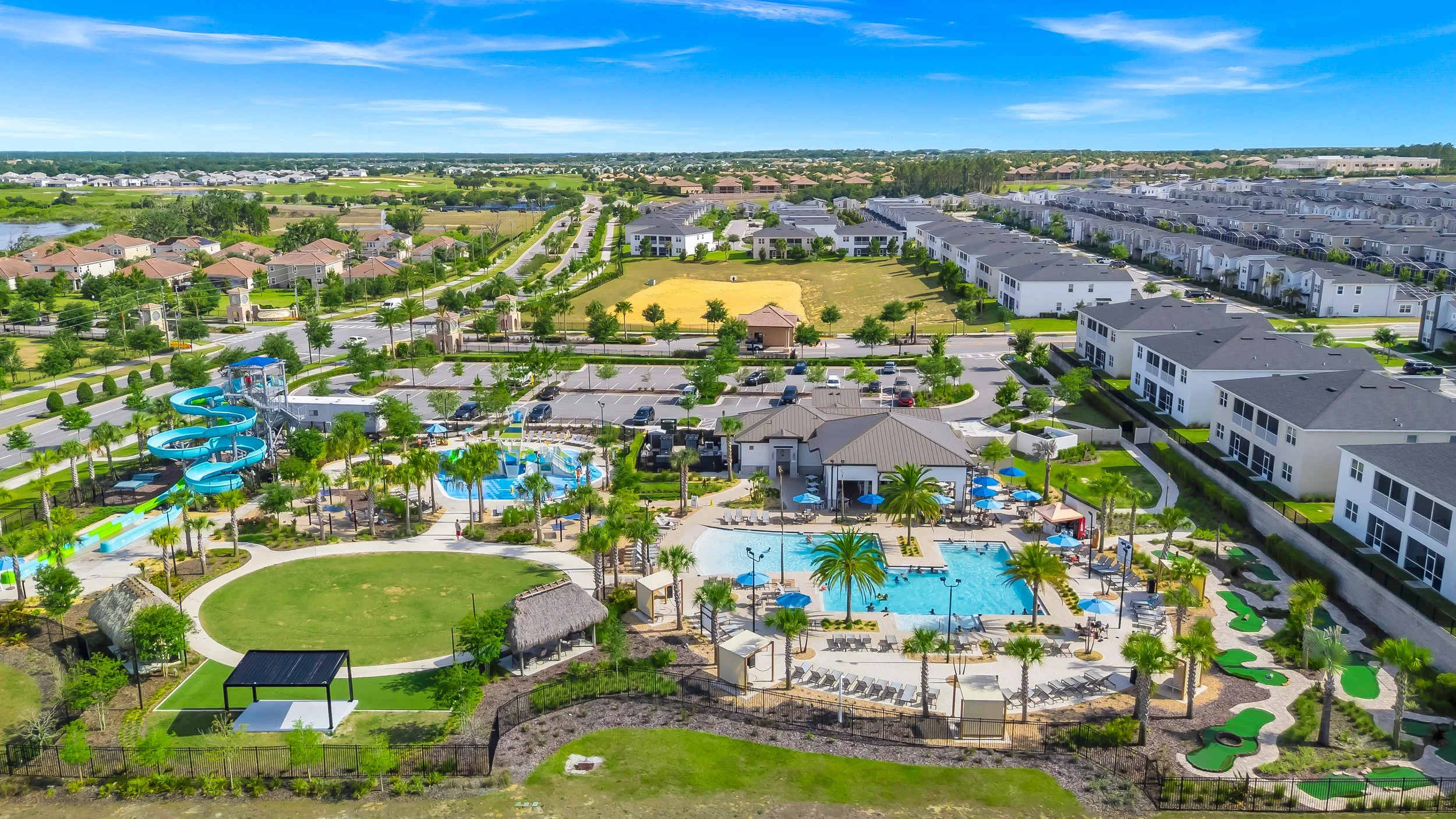 Aerial view of a large residential community with a swimming pool, water slides, and recreational areas, surrounded by houses and green landscapes under a clear blue sky.