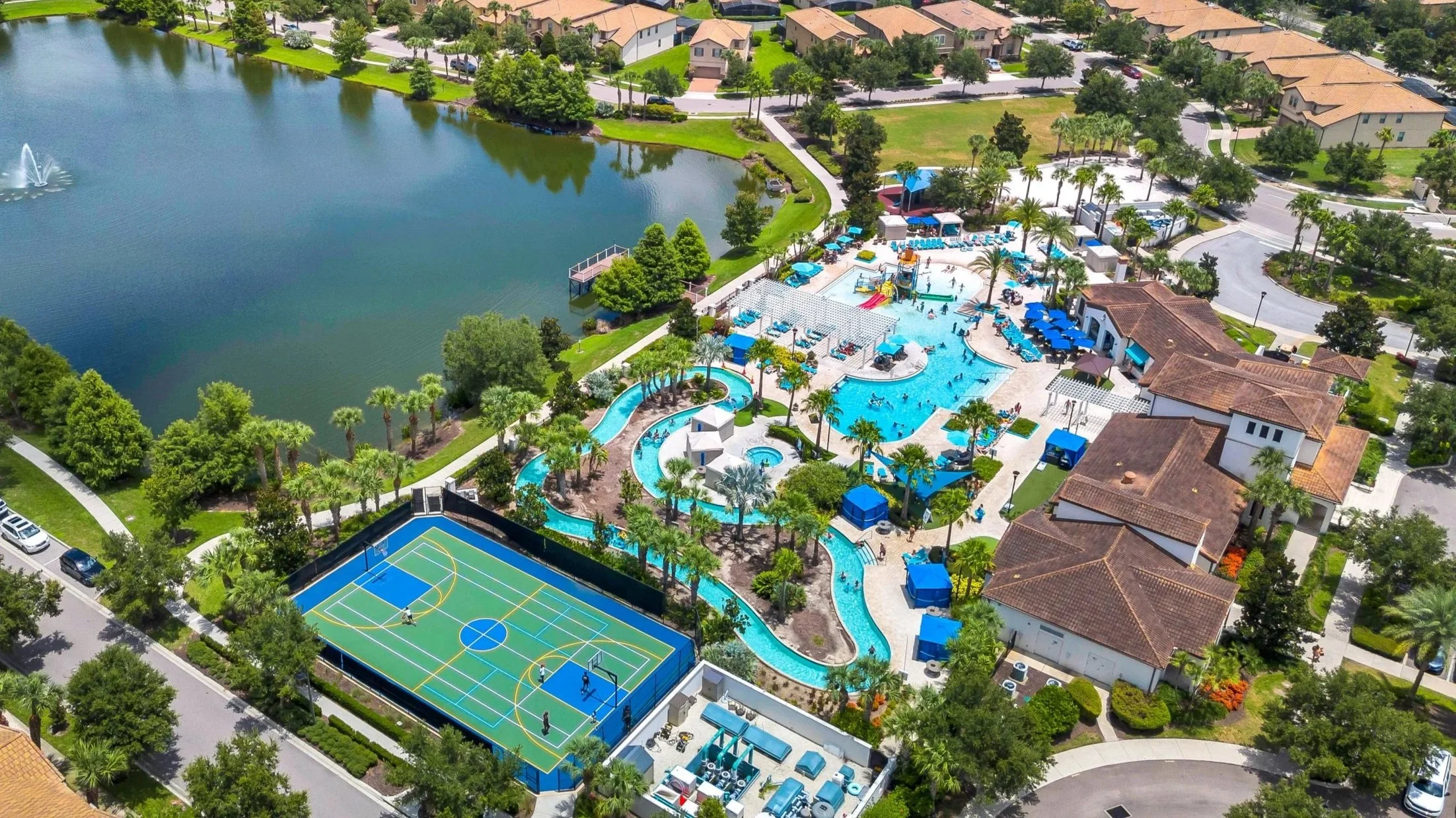 Aerial view of a residential community featuring a large lake with a fountain, a swimming pool area with water slides, and a tennis court, surrounded by lush green trees and houses with tan roofs.