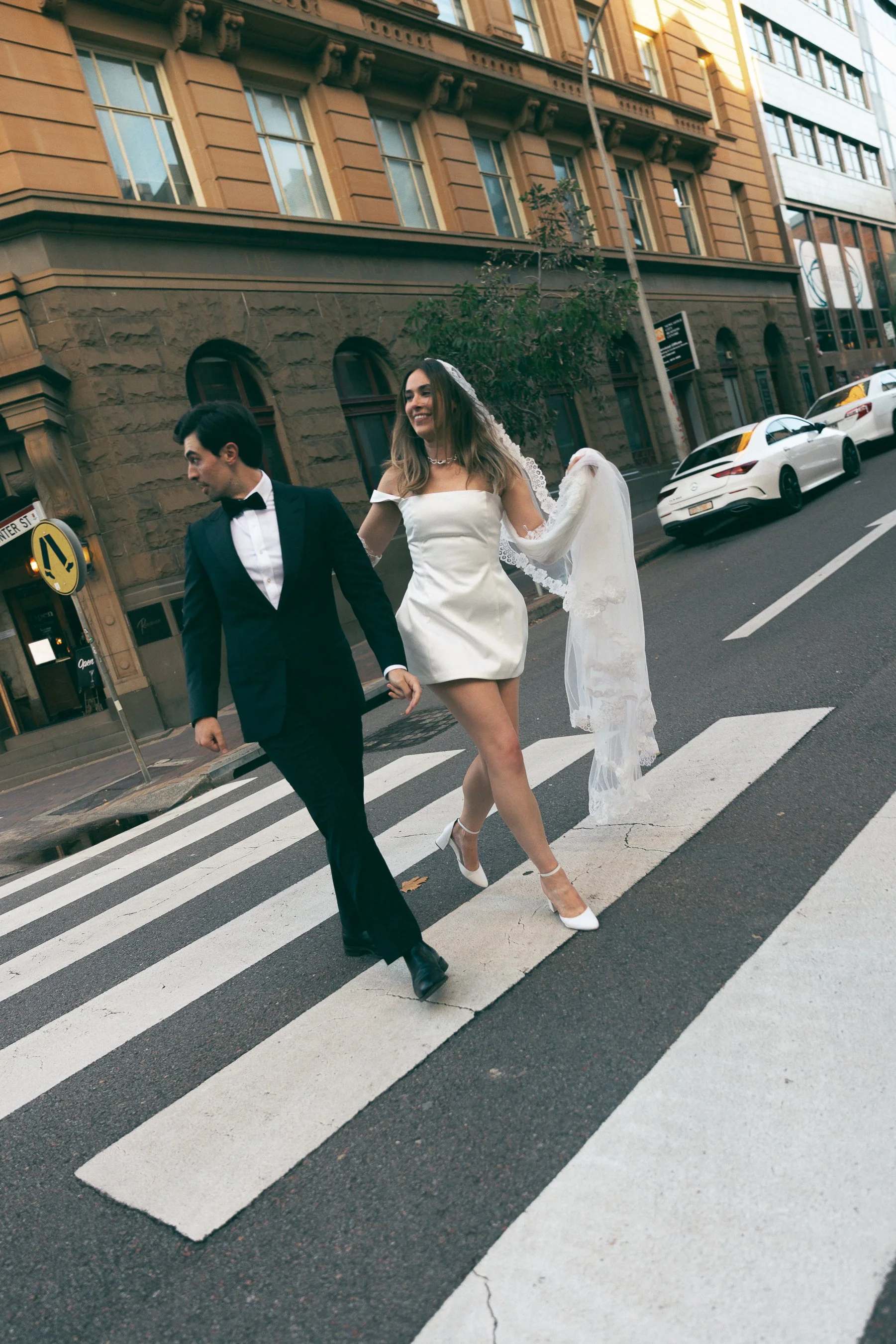 A newlywed couple holding hands while crossing a city street at a crosswalk. The groom is wearing a black tuxedo with a bow tie, and the bride is dressed in a white off-shoulder wedding dress with a veil, high heels, and a necklace. They are smiling 