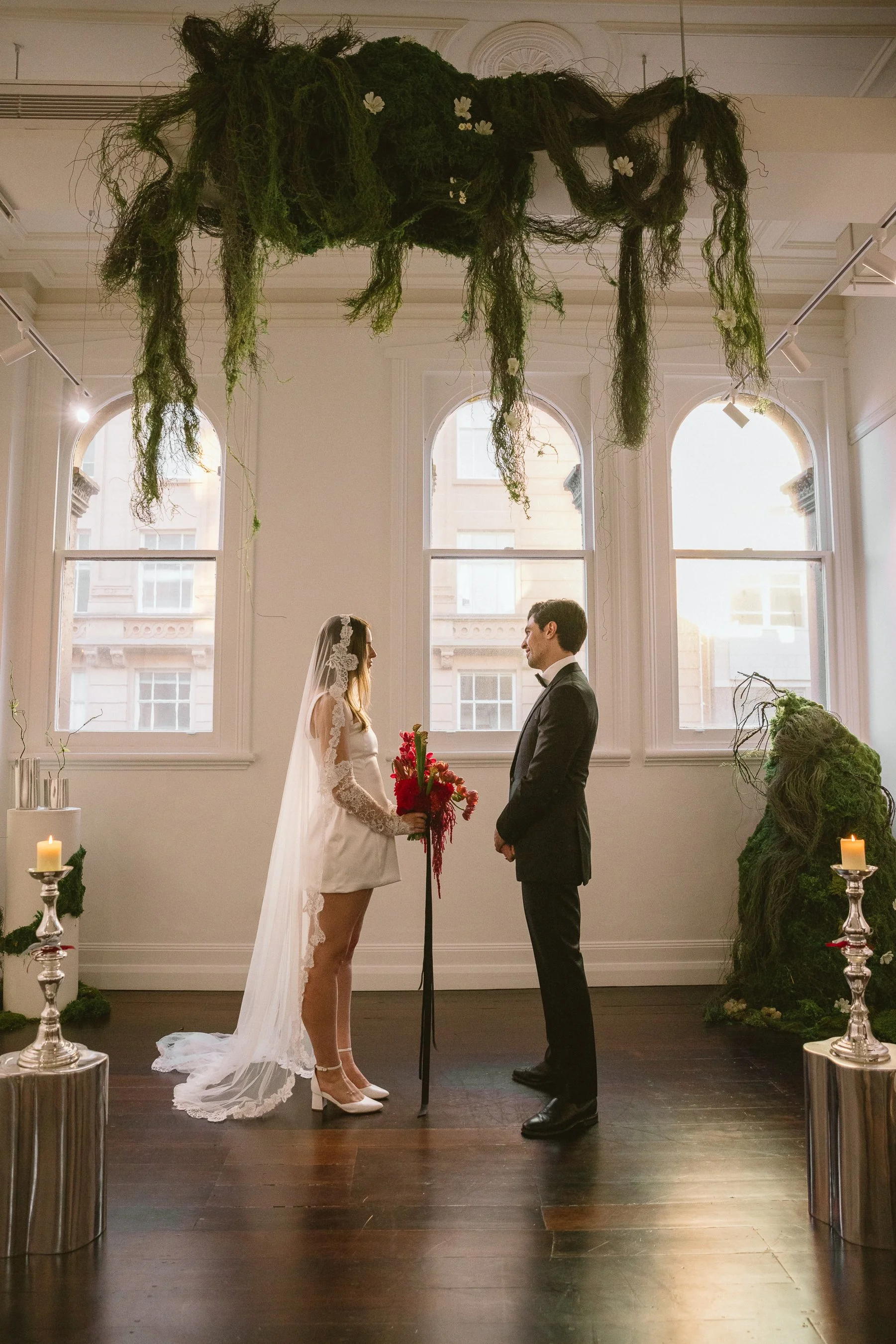 A bride and groom are standing face to face in a room with three large windows and natural light. The bride is wearing a short white bridal dress with lace sleeves and a veil, holding a bouquet of red flowers. The groom is dressed in a black tuxedo. 