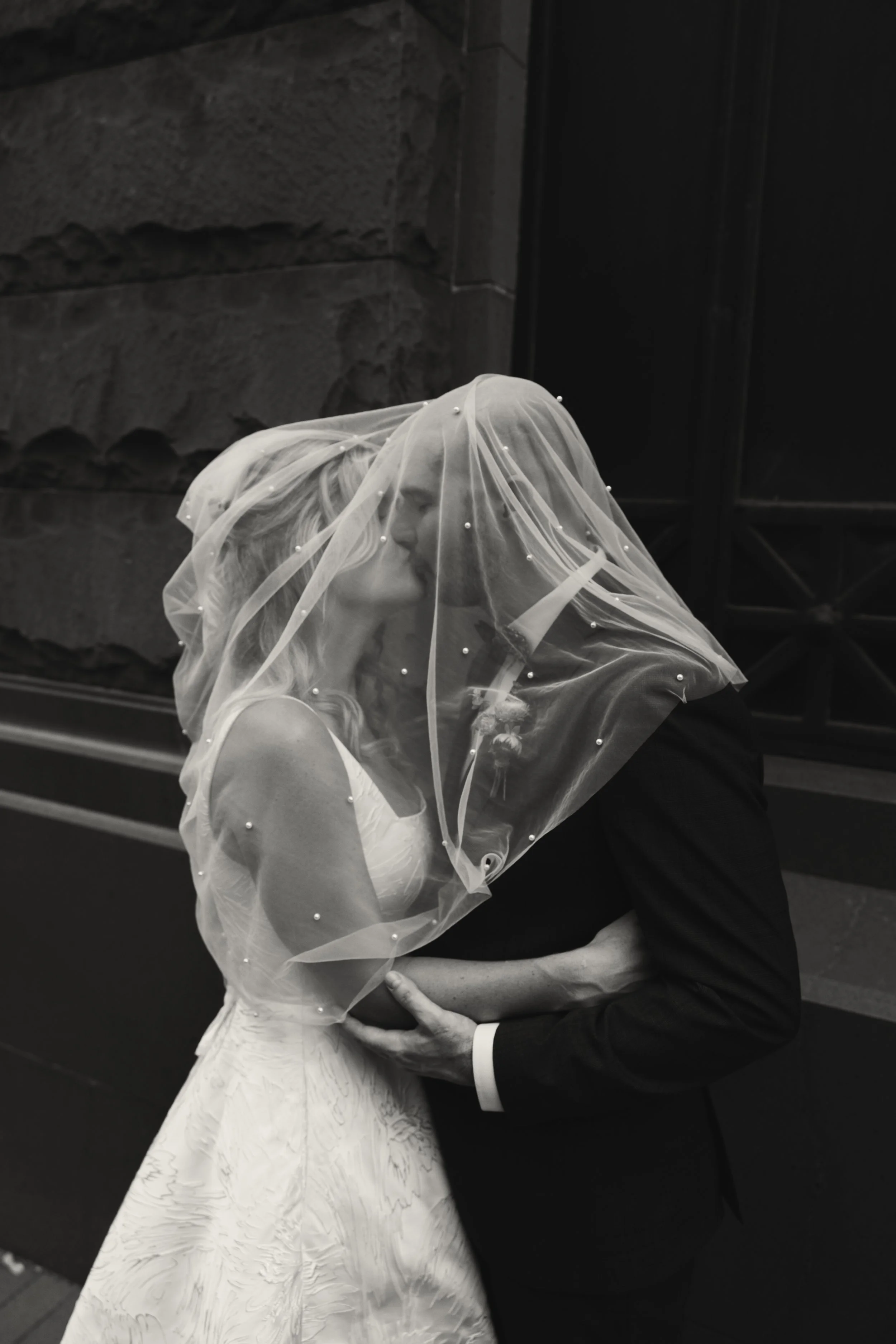 A black-and-white photo of a bride and groom sharing a kiss under a sheer veil decorated with small pearls, standing outdoors in front of a stone wall.