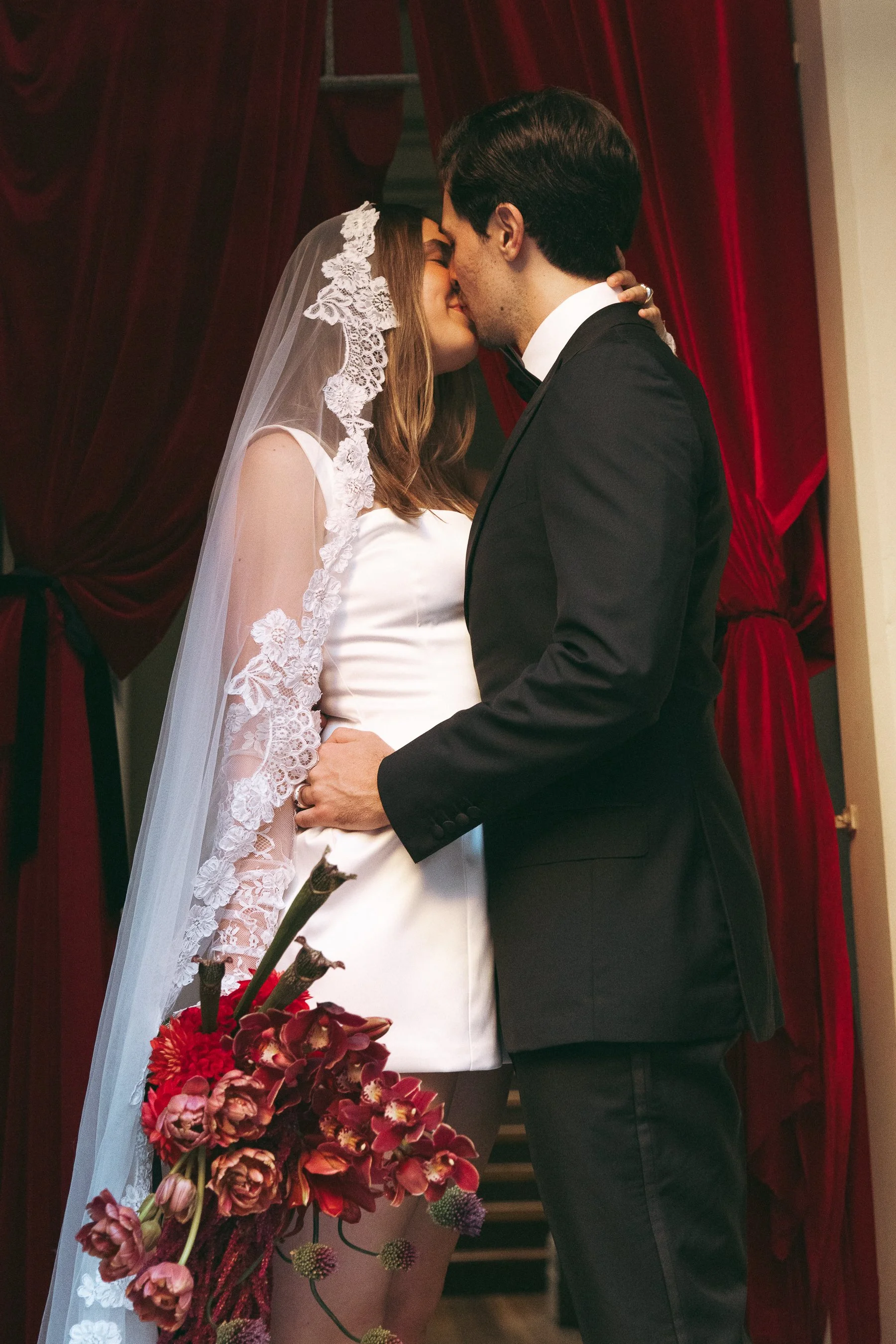 A bride and groom share a kiss during their wedding ceremony, with the bride in a white dress and lace veil holding a bouquet of flowers, and the groom in a black suit, standing in front of red curtains.